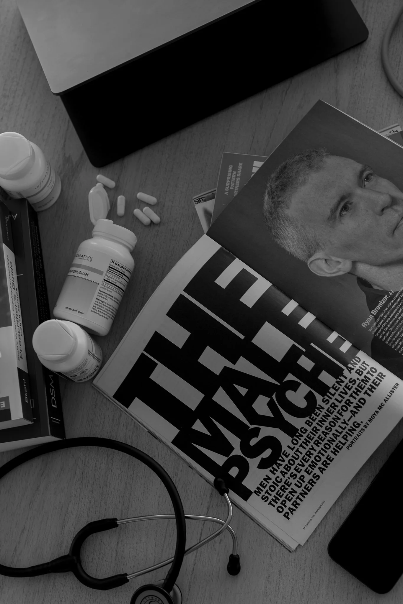 A stethoscope, prescription bottles, and medication pills on a wooden surface next to a magazine and a laptop.