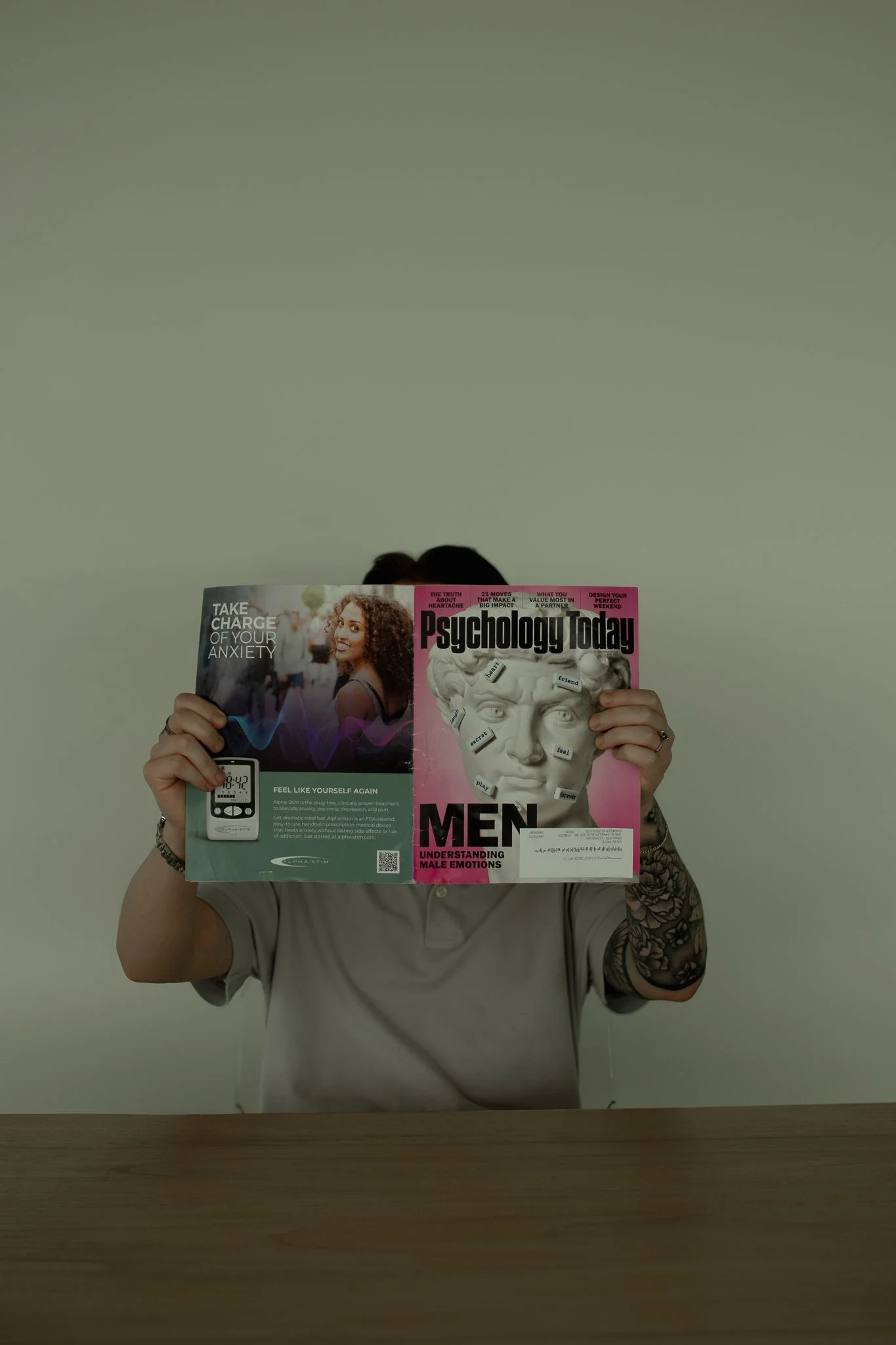 Person holding a magazine titled "Psychology Today" in front of their face, revealing tattoos on their right arm and wearing a beige shirt, with a plain light-colored wall in the background.
