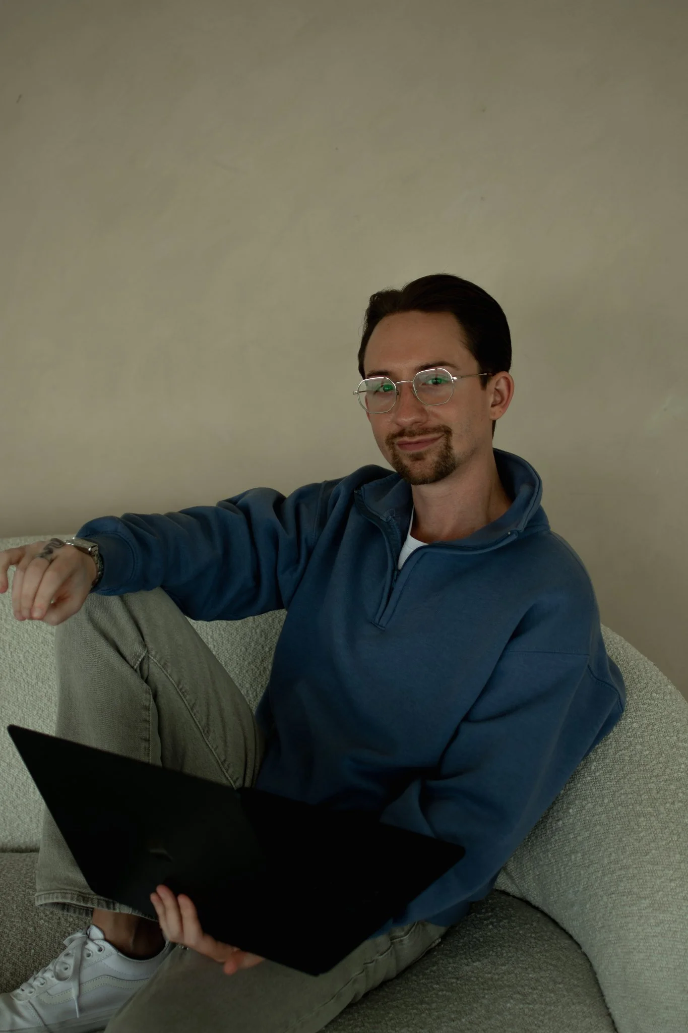 A young man with glasses and a goatee sitting on a grey couch, working on a laptop, wearing a blue jacket, light grey pants, white sneakers, and a watch, with a beige wall in the background.