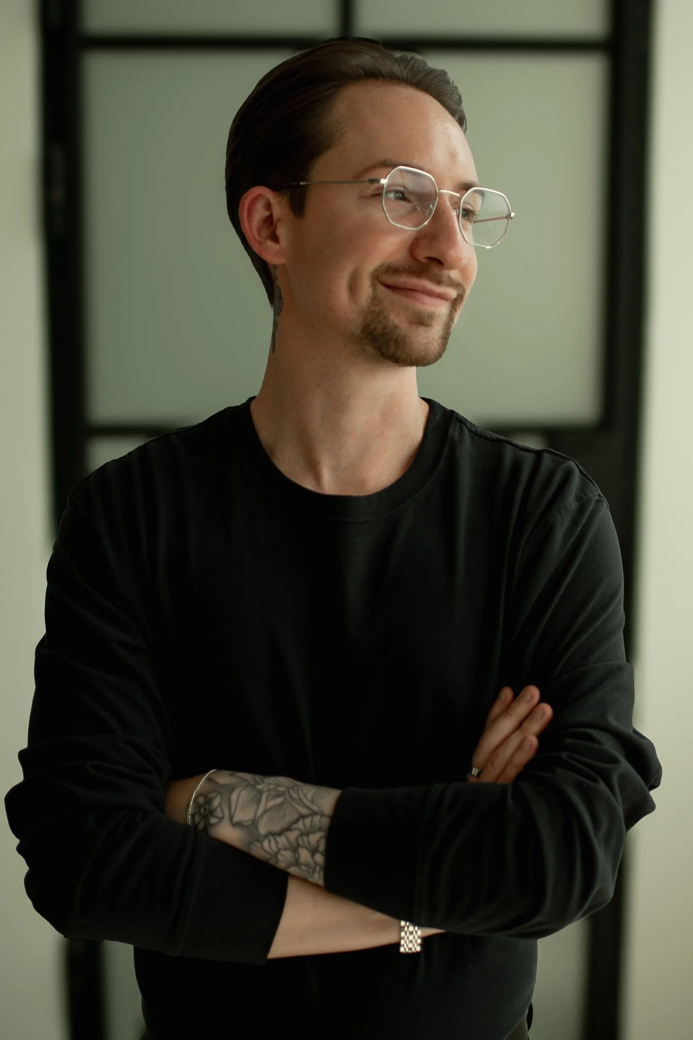 A man with brown hair, glasses, and a goatee smiling and crossing his arms, wearing a black long-sleeve shirt, with a tattooed arm, standing in front of a dark framed background.