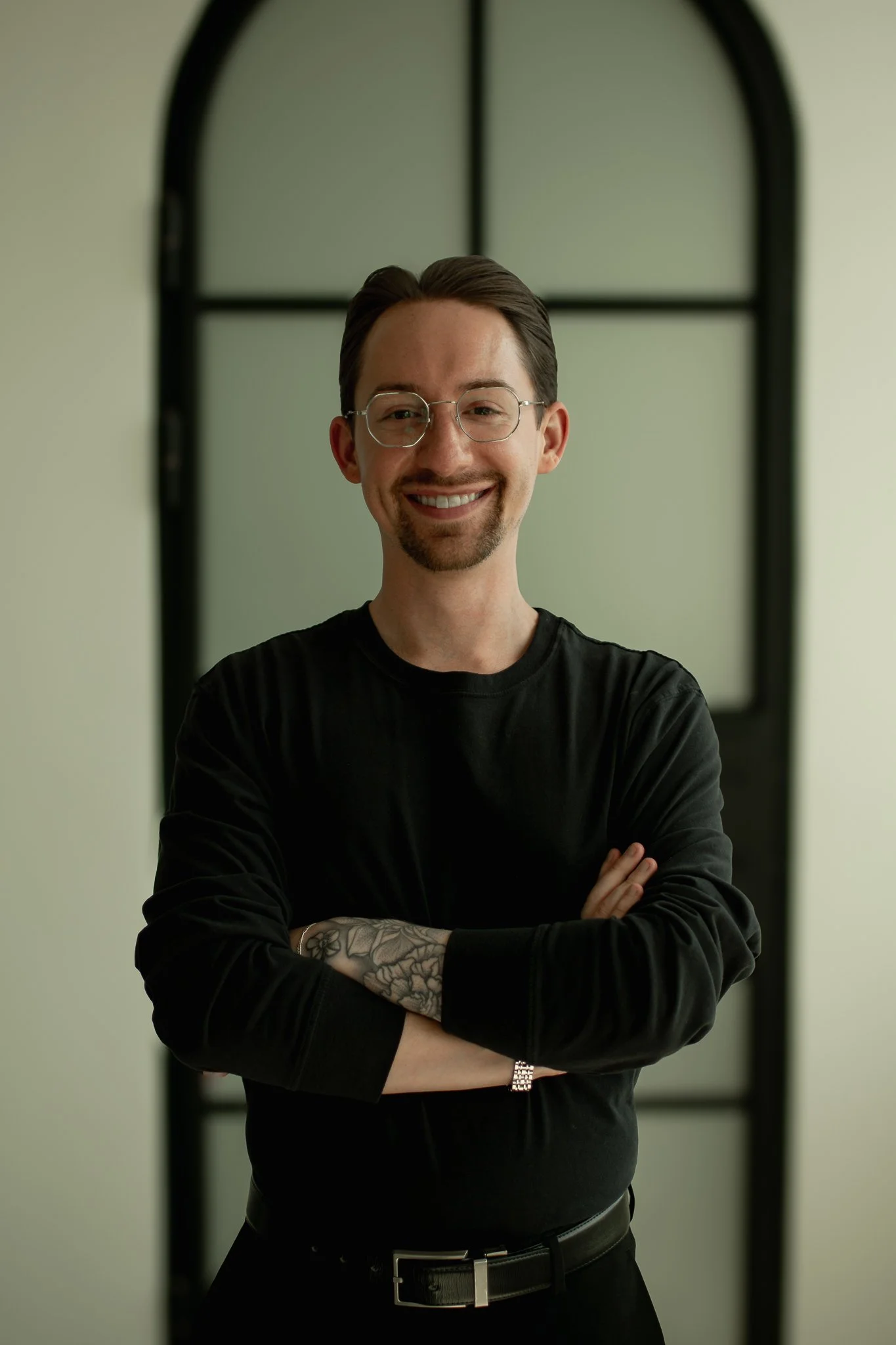 A smiling man with dark hair, glasses, and a beard, wearing a black shirt and silver watch, standing with crossed arms in front of a mirror with a black frame.