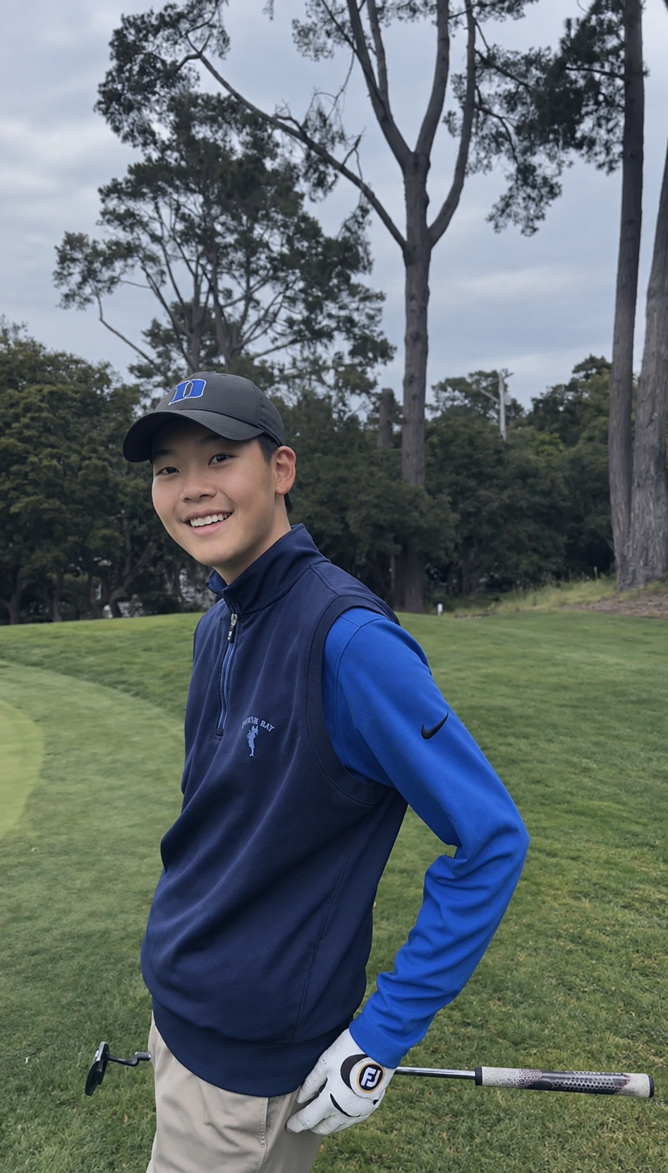A young man smiling on a golf course, holding a golf club in his right hand. He is wearing a black cap, a blue and black Nike vest with a logo that reads "Golf Bay," and beige pants. The background features green grass, tall trees, and overcast sky.