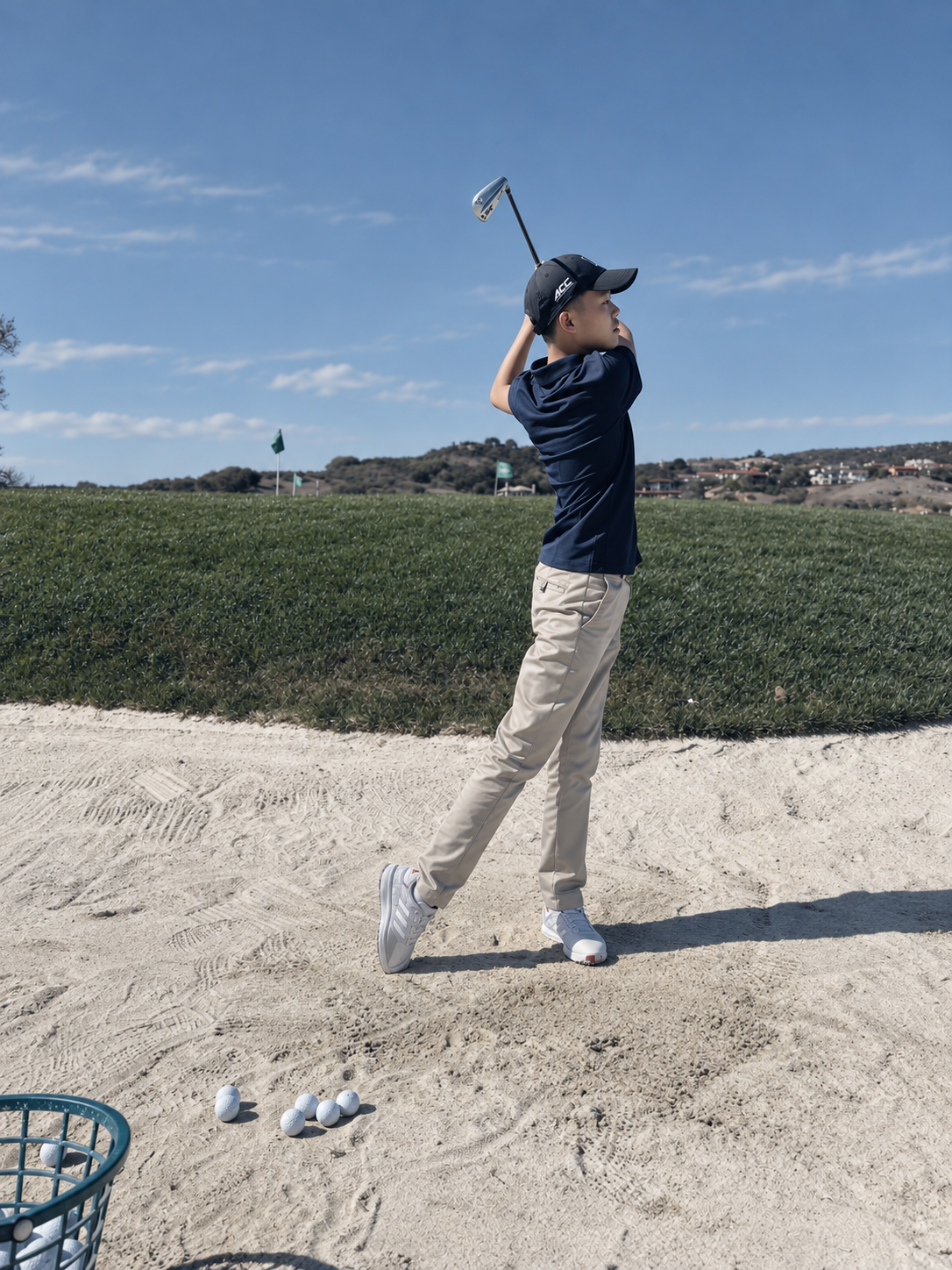 A boy playing golf, standing on a sand bunker, holding a golf club, with golf balls and a basket nearby, on a golf course with a grassy field and a blue sky.