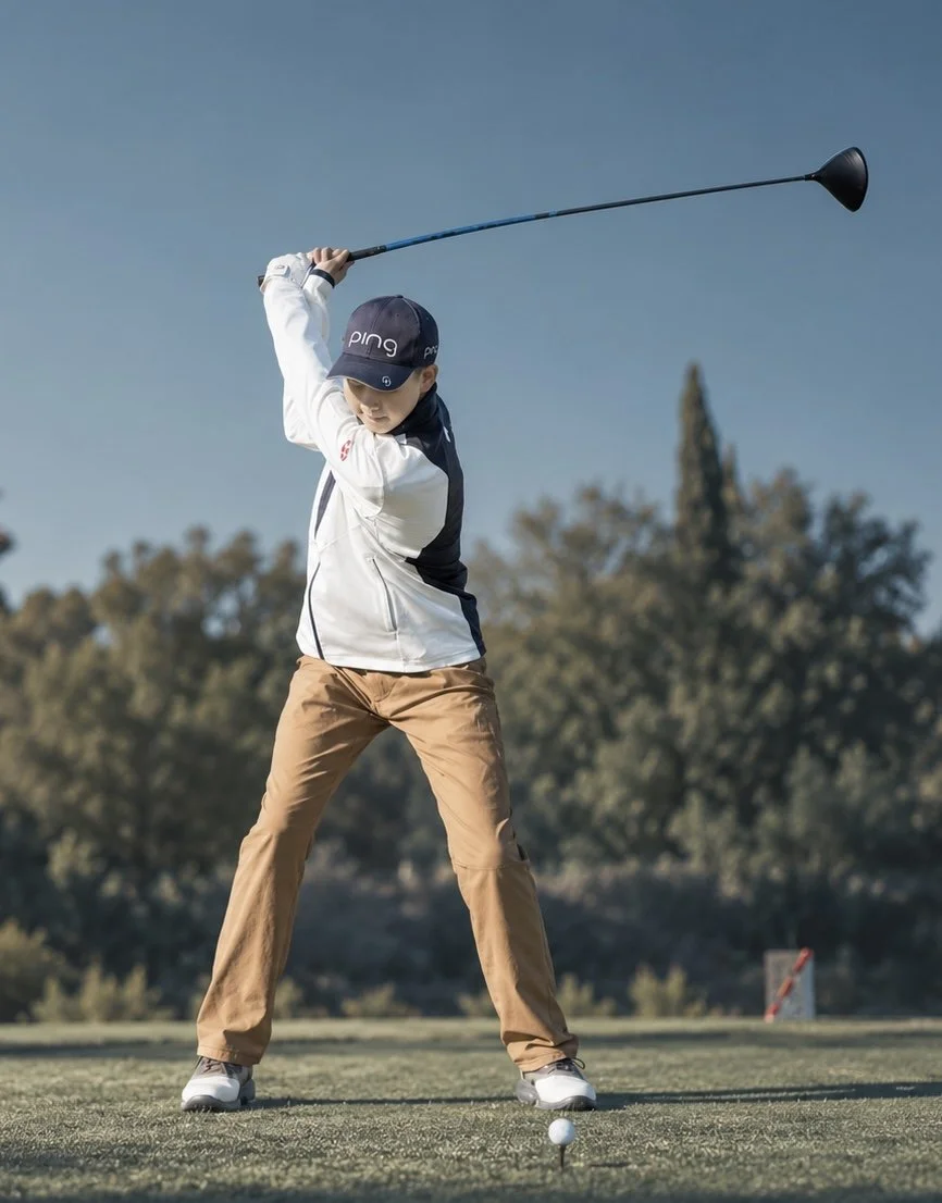 Young boy wearing a golf cap and jacket swings a golf club on a golf course, preparing to hit a golf ball.