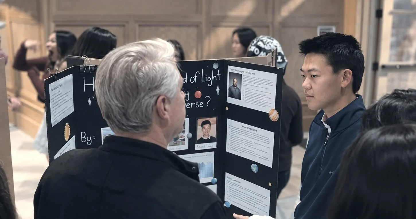 An older man and a young man engaging in a science fair discussion at a presentation board titled 'Light of Light in the Universe?' with various texts, images, and graphs, while other people are visible in the background.