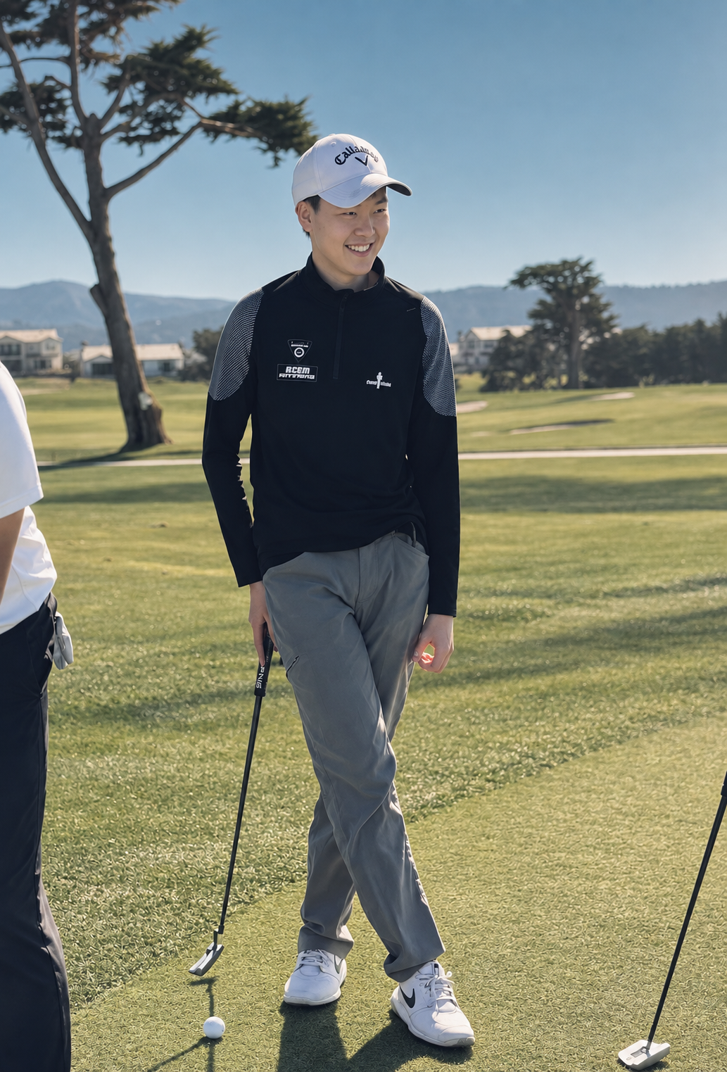 A young man with light skin and short hair standing on a golf course, smiling. He is wearing a white Callaway cap, a black and gray athletic jacket, gray pants, and white Nike golf shoes, holding a golf club.