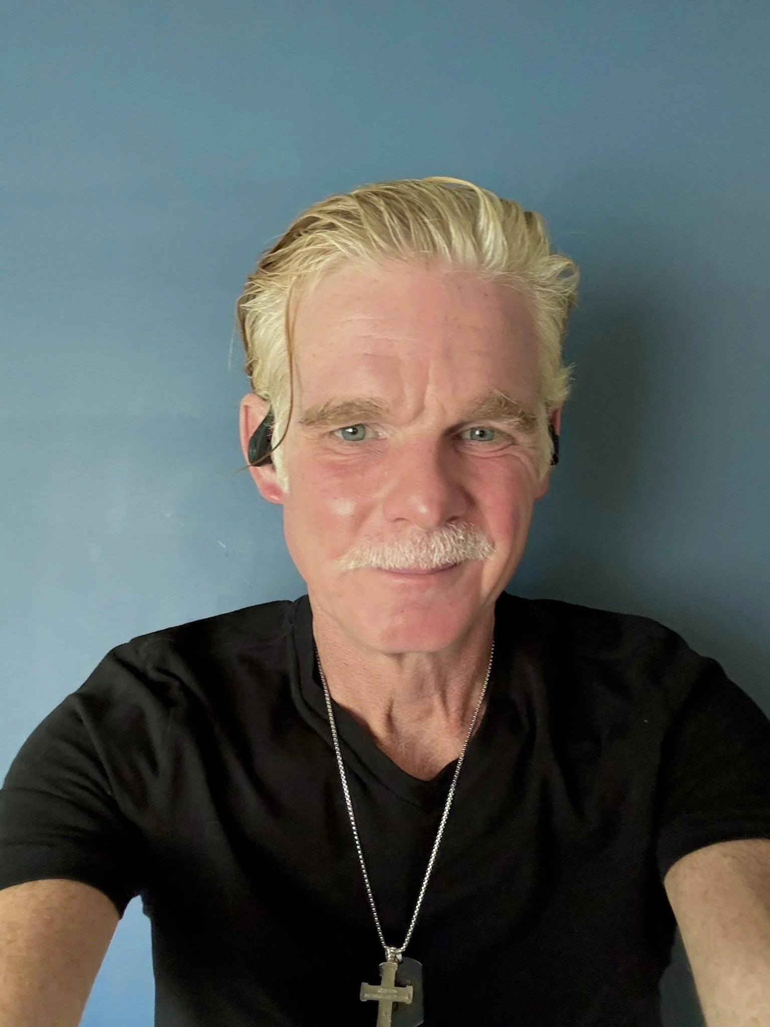 Close-up of a smiling middle-aged man with blond hair, a mustache, and a beard, wearing a black shirt and a cross necklace, taking a selfie against a blue background.