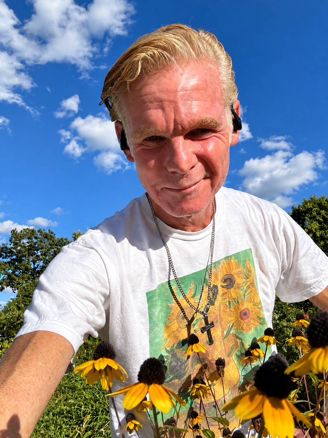 A man with blonde hair taking a selfie outdoors in a garden with yellow flowers and trees. He is wearing earbuds, necklaces with crosses, and a white T-shirt featuring sunflowers and a woman wearing a sun hat. The sky is blue with scattered white clouds.