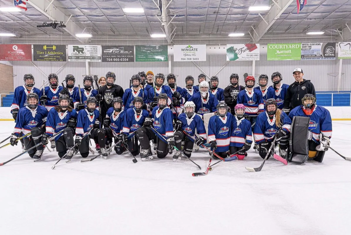 A youth ice hockey team in blue jerseys posing for a group photo on the ice rink, with coaches and team members all wearing helmets and hockey gear, inside an indoor ice rink.