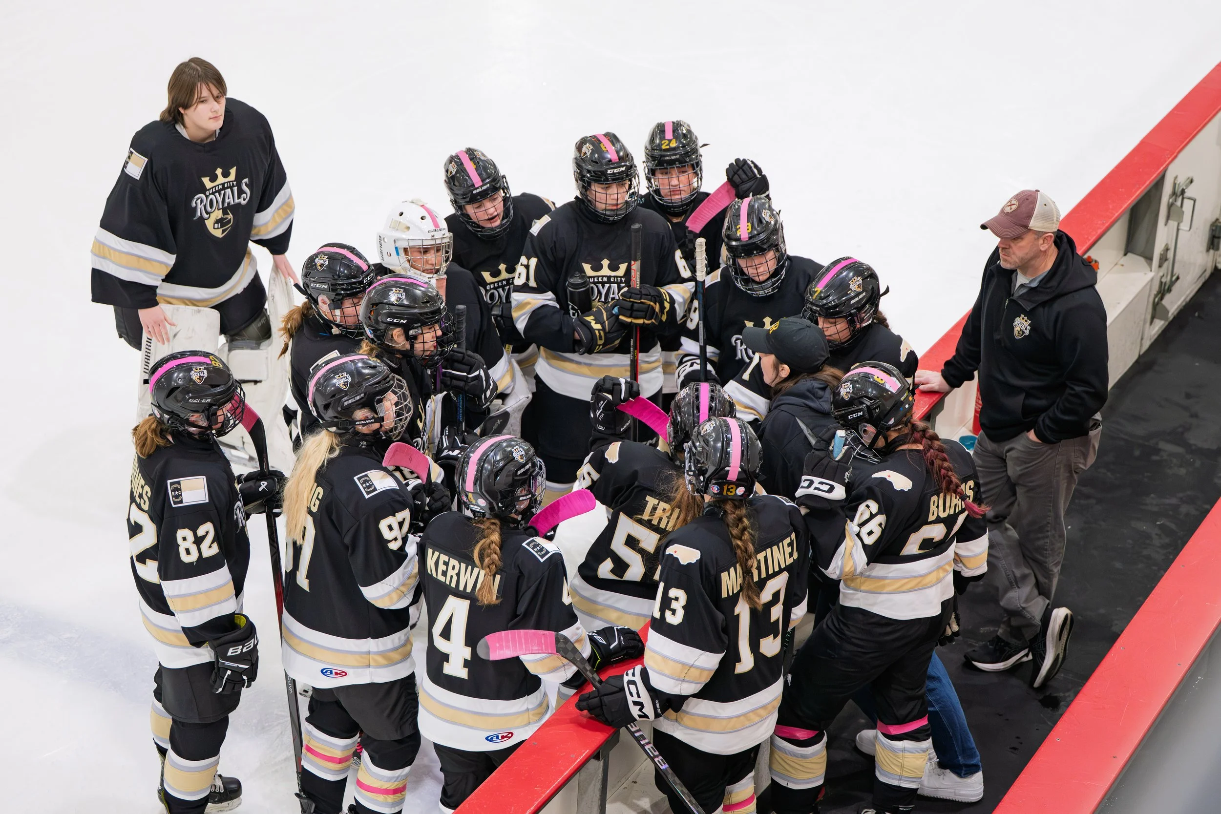 A women's ice hockey team is gathered on the bench during a game. They are wearing black jerseys with gold and white details, and pink tape on their sticks and helmets. The team appears to be listening to their coach, who is dressed in a black jacket and cap, speaking to them.