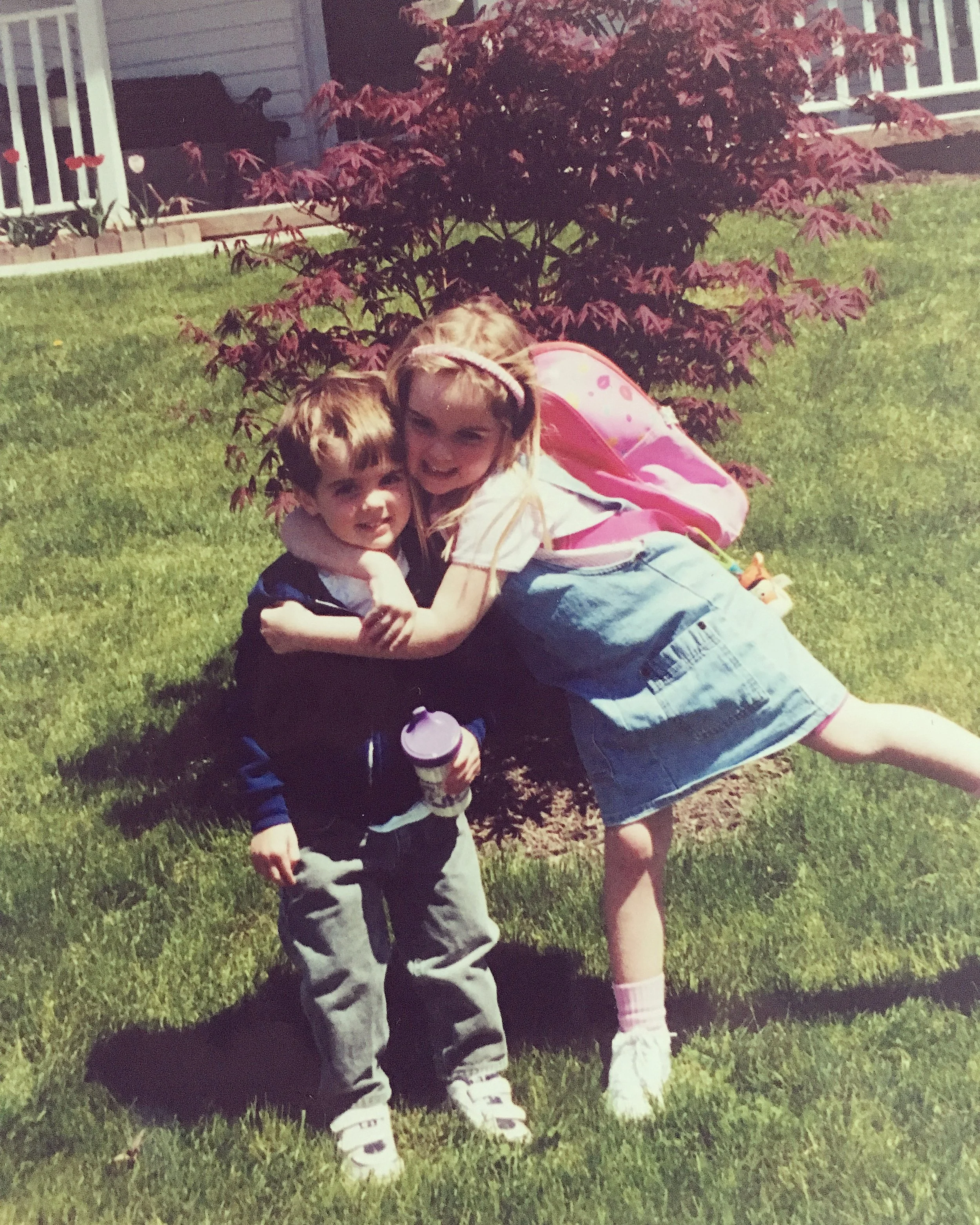 Two children hugging on a grassy lawn, with a small red-leafed tree and a house in the background.