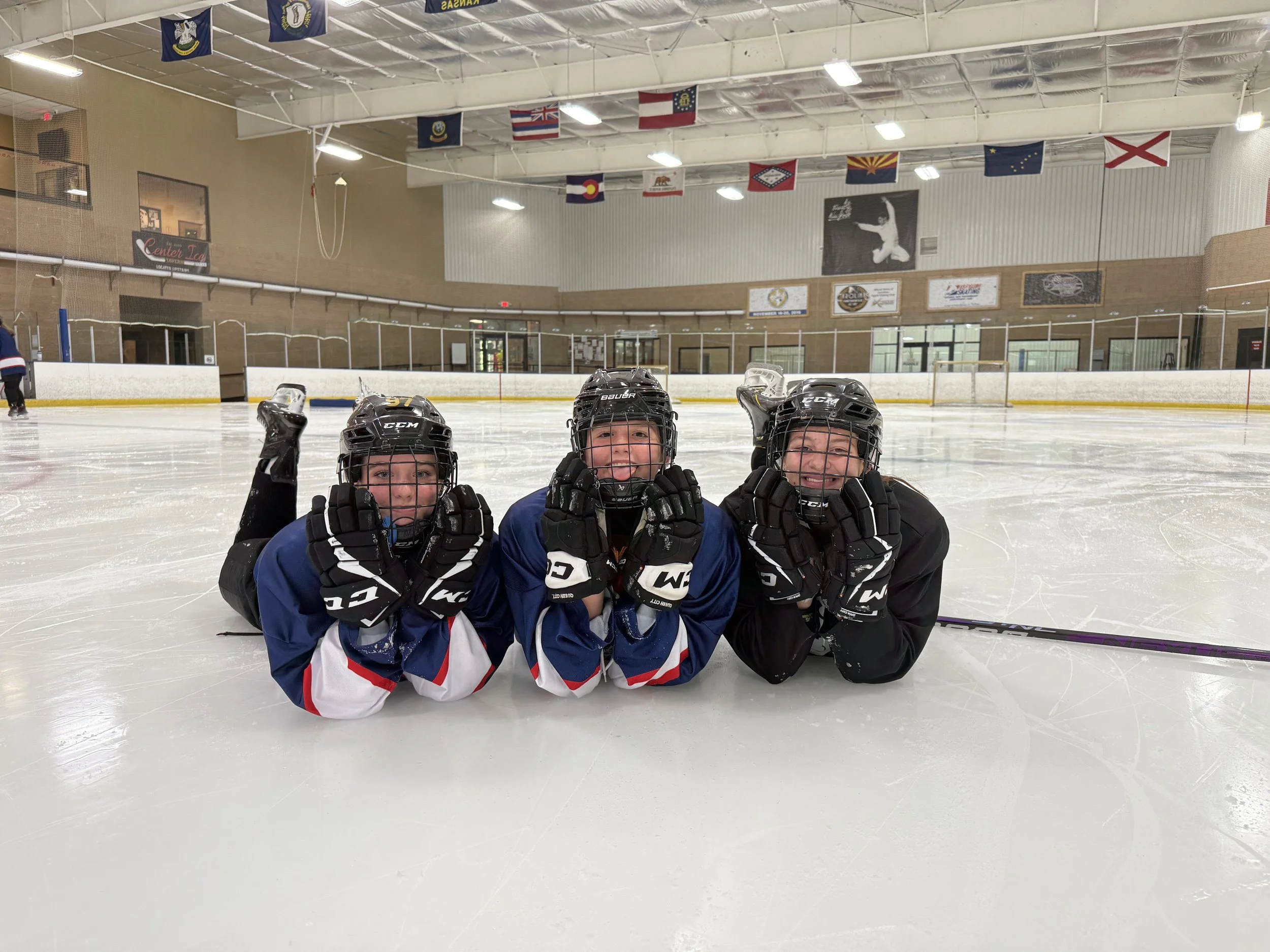 Three children in hockey gear laying on the ice rink with helmets and gloves, smiling, inside an indoor ice hockey rink.