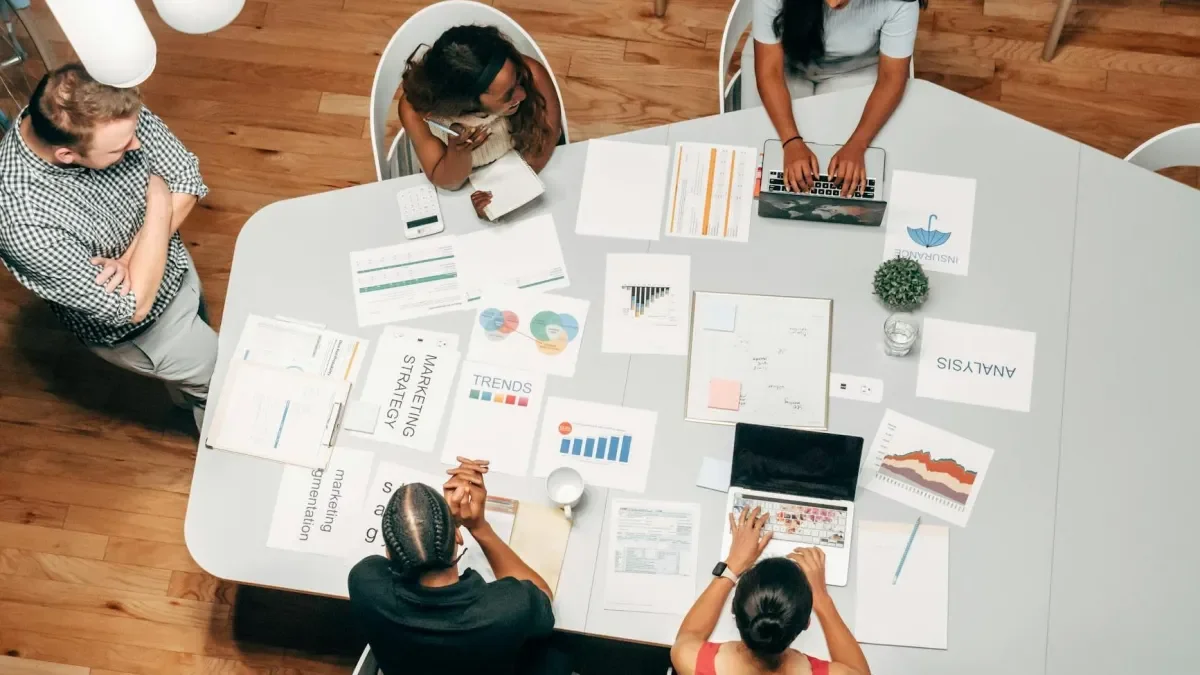 A group of people working on a business meeting around a large white table with charts, laptops, notebooks, and documents.