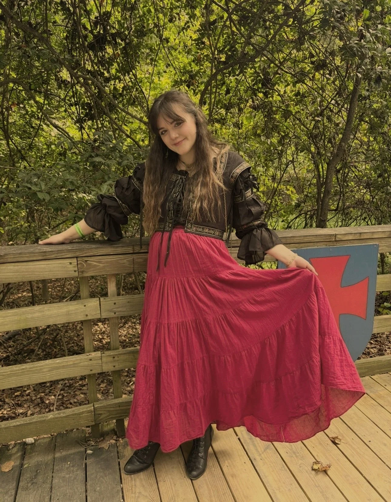 A woman in a black long-sleeved dress with ruffled sleeves and a red skirt, standing on a wooden deck with a forest in the background. She is smiling slightly, holding the skirt with one hand, and has long, wavy brown hair.