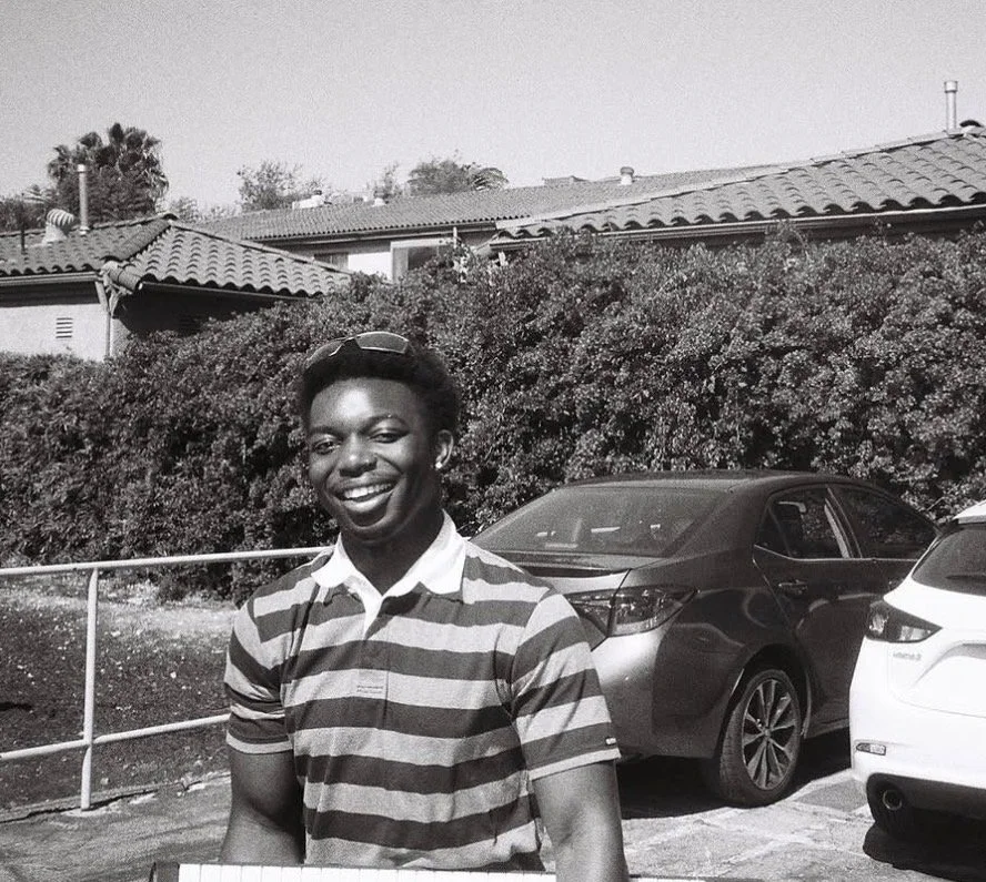 A young man smiling outdoors, wearing a striped polo shirt, sunglasses on his head, in a parking lot with cars and bushes, with houses in the background.