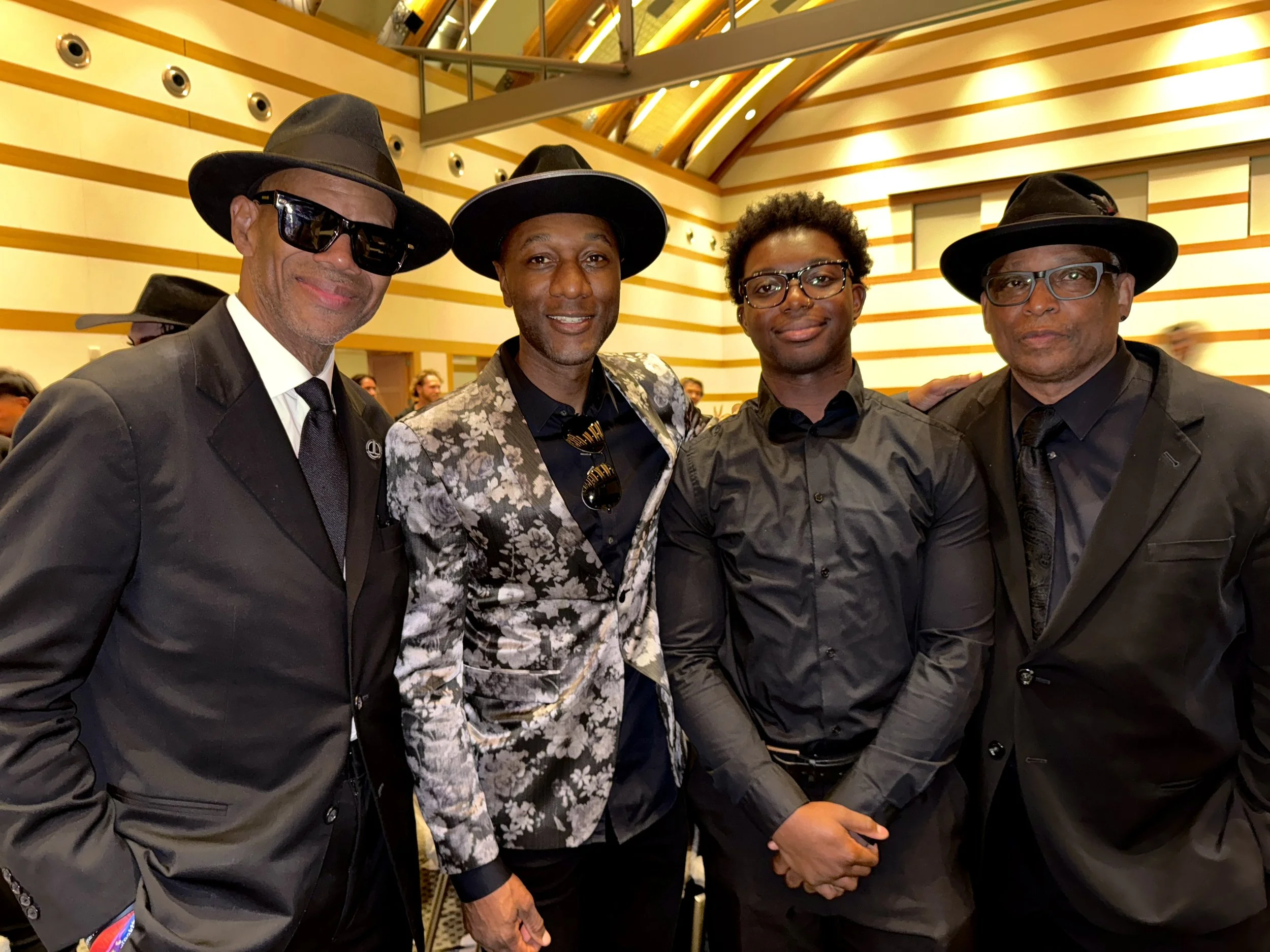Four men dressed in black suits, with three wearing black cowboy hats and one wearing glasses, standing together indoors in a modern building with yellow and orange walls.
