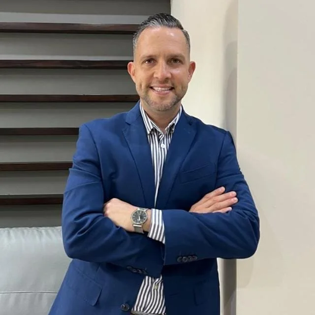 Hombre sonriendo con blazer azul y camisa a rayas, de pie en un interior.