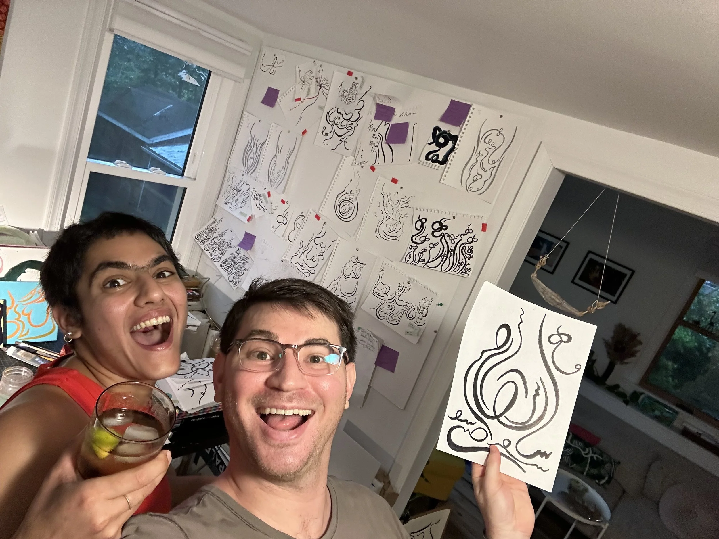 Two smiling people celebrating, holding a glass of wine and a piece of artwork with abstract black line designs on white paper. The background displays numerous similar artworks taped to a wall in a room with a window showing an outdoor scene.