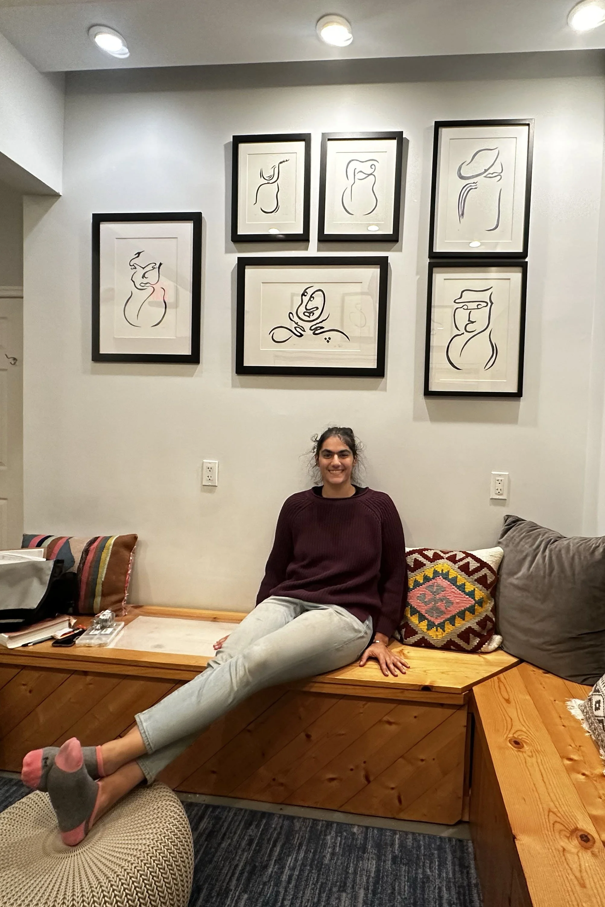 A woman in a burgundy sweater and gray pants sitting on a wooden bench with colorful pillows . Framed abstract artwork hangs on the white wall behind her, and various items are on a table to her left, with a patterned carpet and other furniture around.