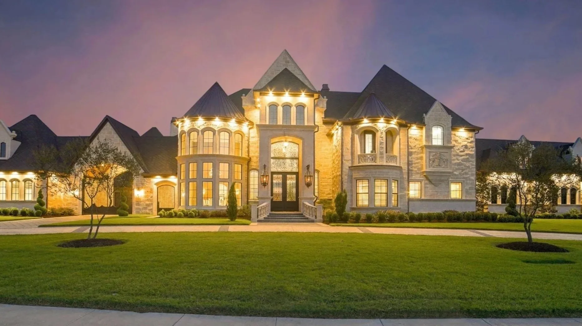 Large, elegant mansion with stone and stucco exterior, multiple turrets, arched windows, and front steps illuminated at dusk, surrounded by a manicured lawn and small trees.