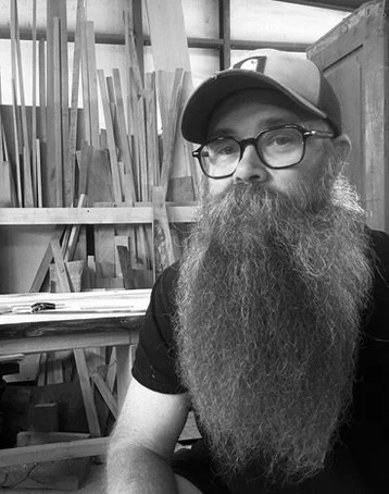 A man with glasses and a cap sitting in a woodworking shop with various wooden planks behind him.