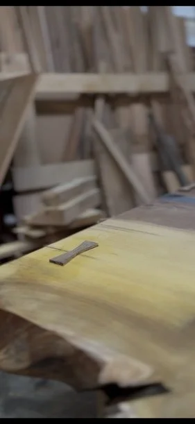 Close-up of a wooden workbench with a small chisel and a broad plank of wood in a woodworking shop filled with various wooden planks and tools in the background.