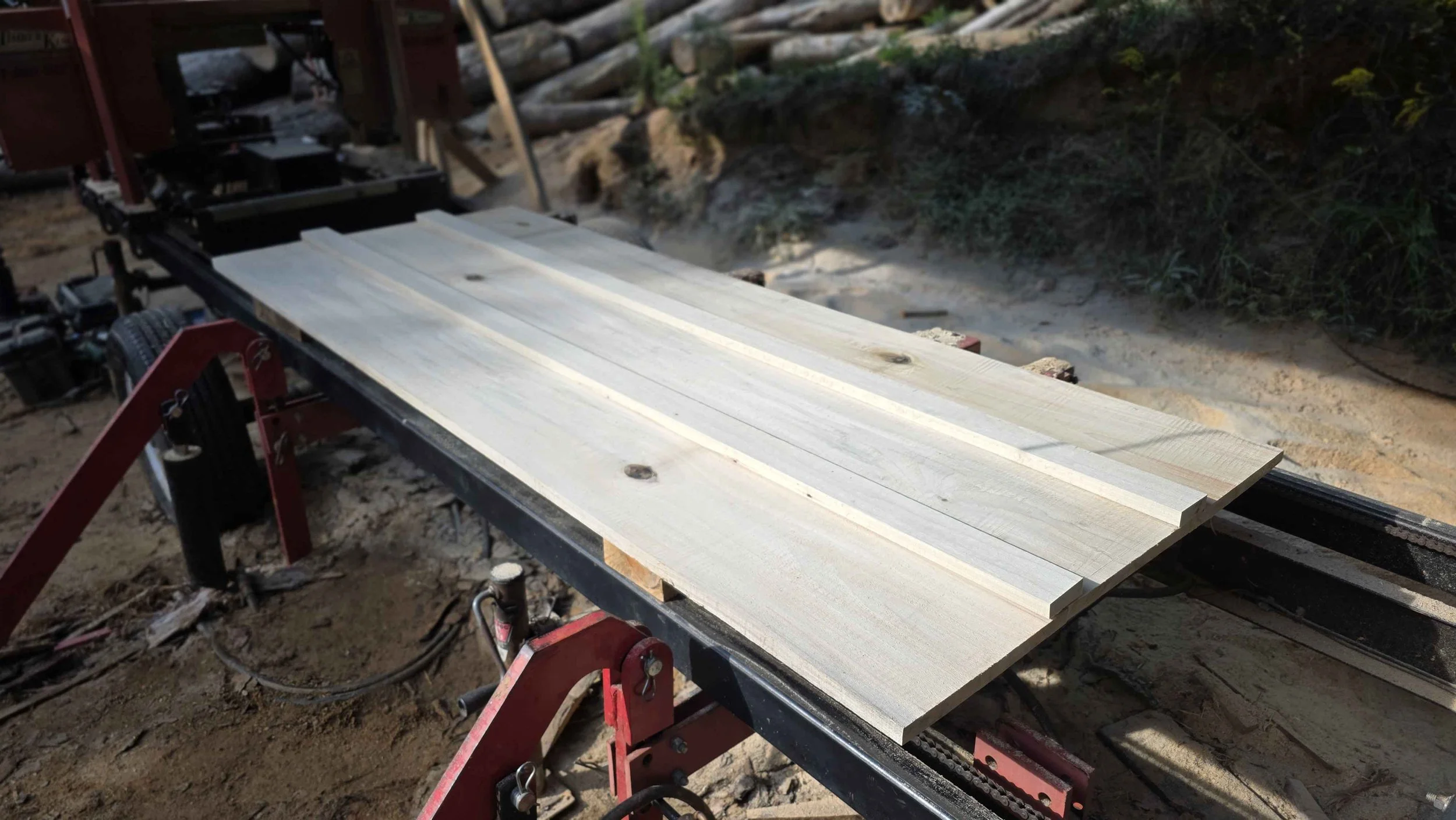 Wood planks on a conveyor or sawmill in an outdoor construction site with soil and logs in the background.