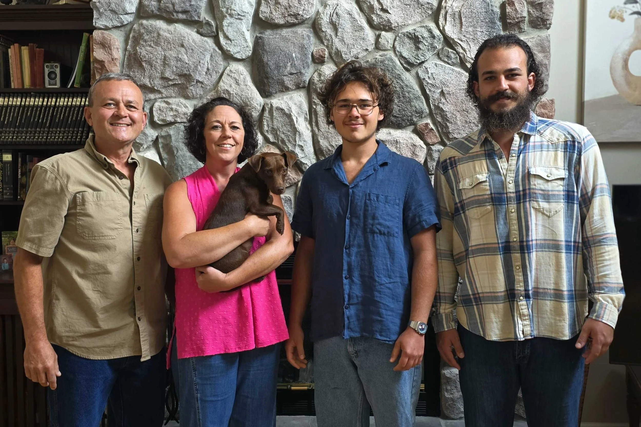 A group of four adults and a young man standing in front of a stone wall. The woman is holding a small brown puppy. There are bookshelves and a fireplace in the background.