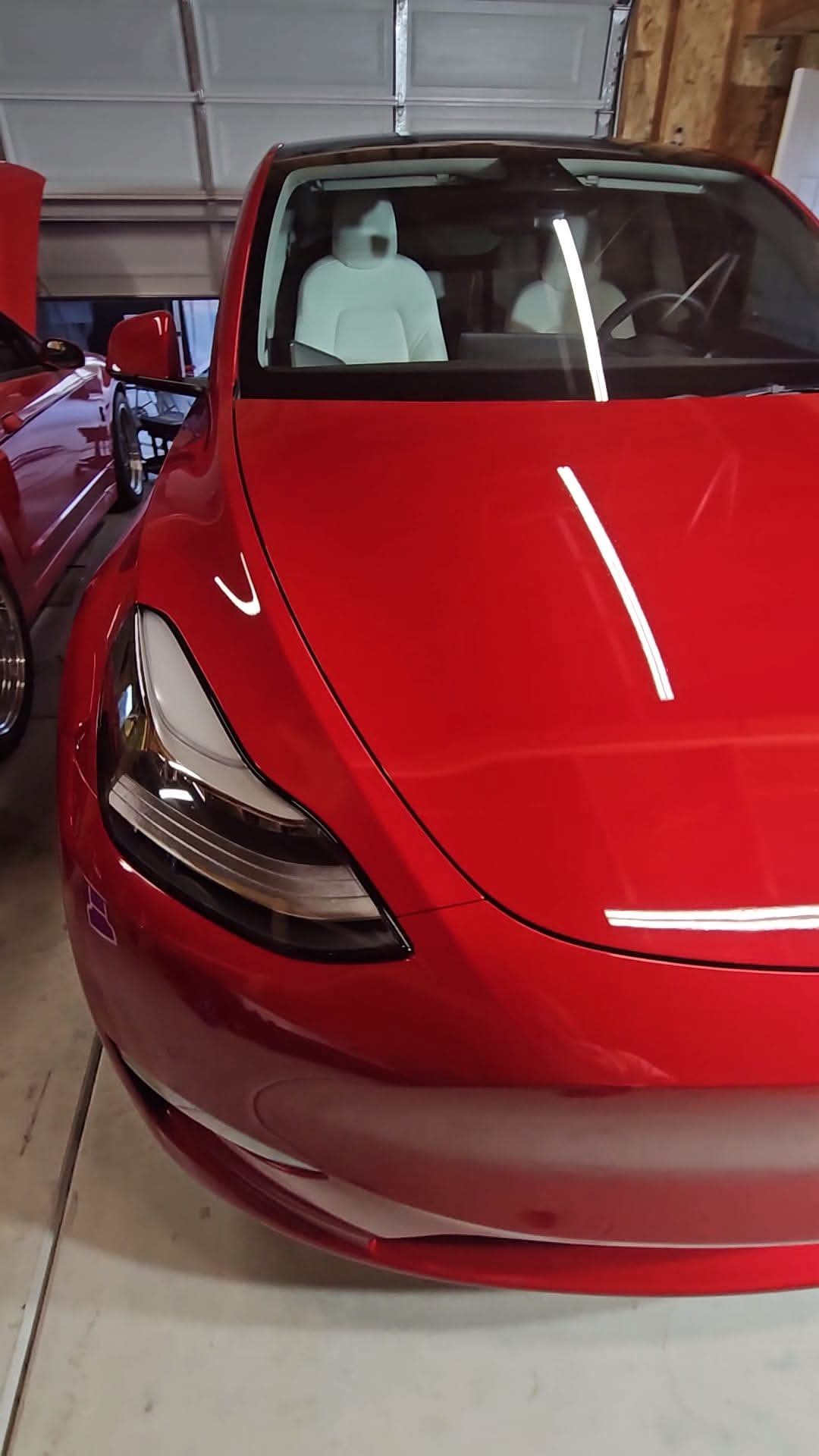 Red sports car parked in a garage next to another car, with a garage door and wooden wall in the background. Paint Correction 