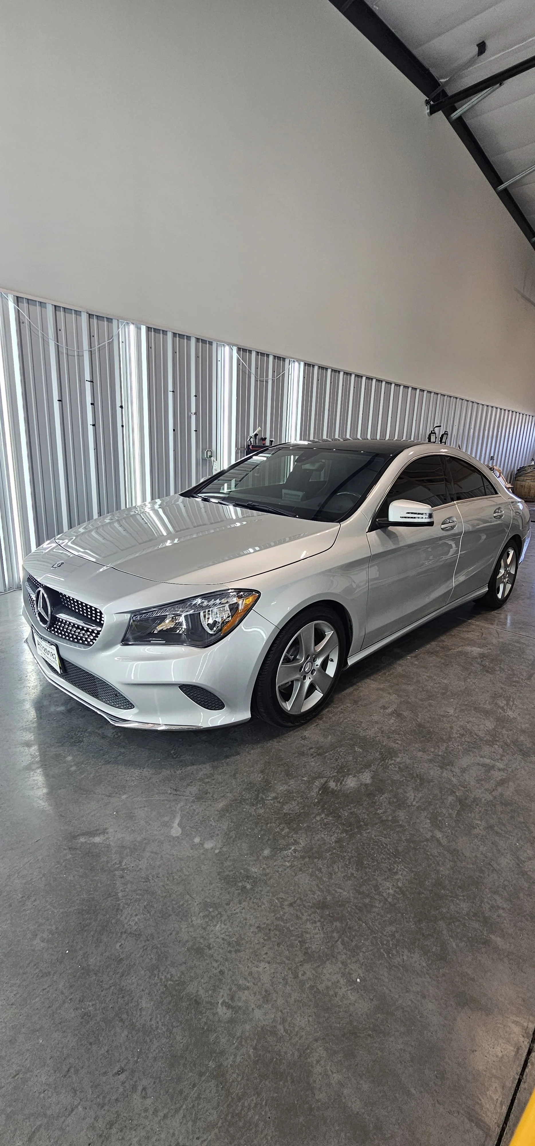 Silver Mercedes-Benz sedan parked inside a garage with a concrete floor and metal wall. The garage has a partially open door, and the car's front view is visible.