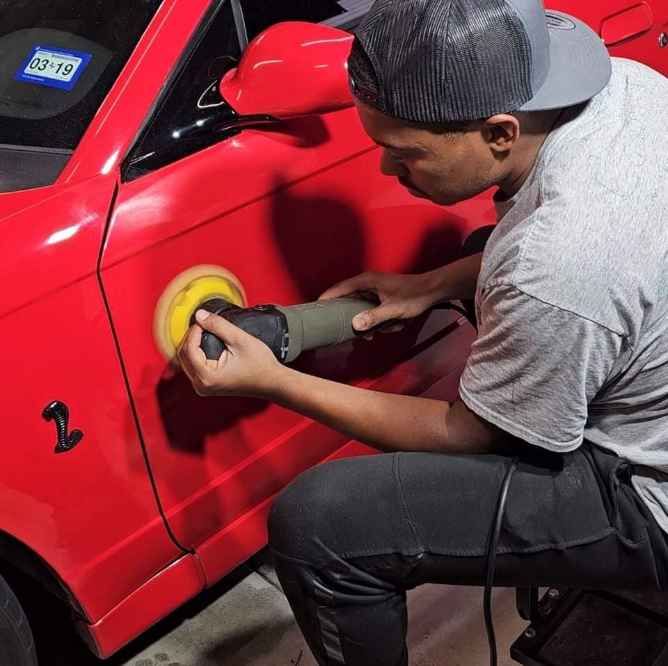 A man polishing or buffing a red sports car with a handheld polishing device inside a garage.