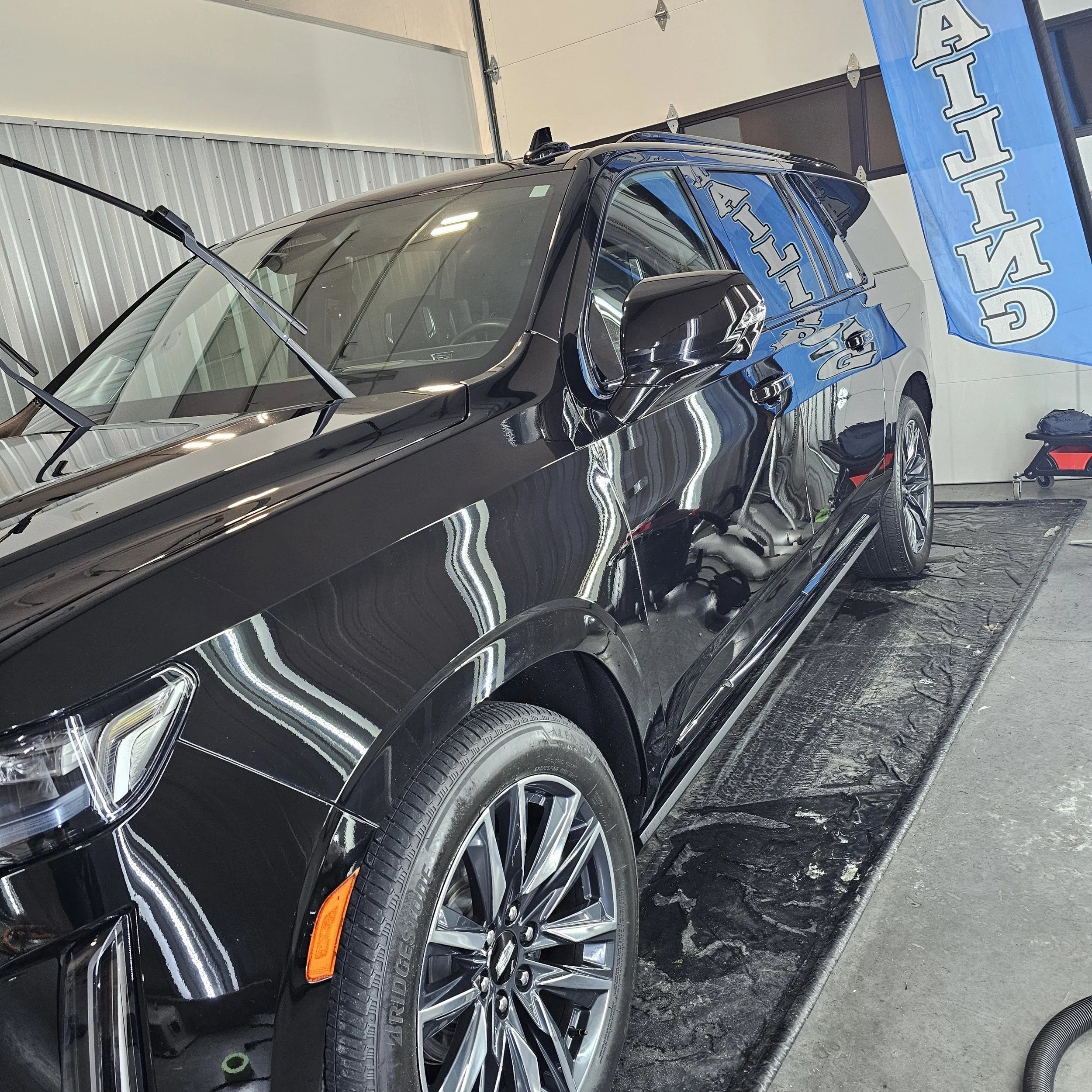 Black SUV parked inside a garage with a large blue banner. The vehicle has shiny black paint, tinted windows, and detailed alloy wheels. The garage has a metal wall, and the floor appears to be slightly wet or polished. Ceramic coating Gadsden, AL