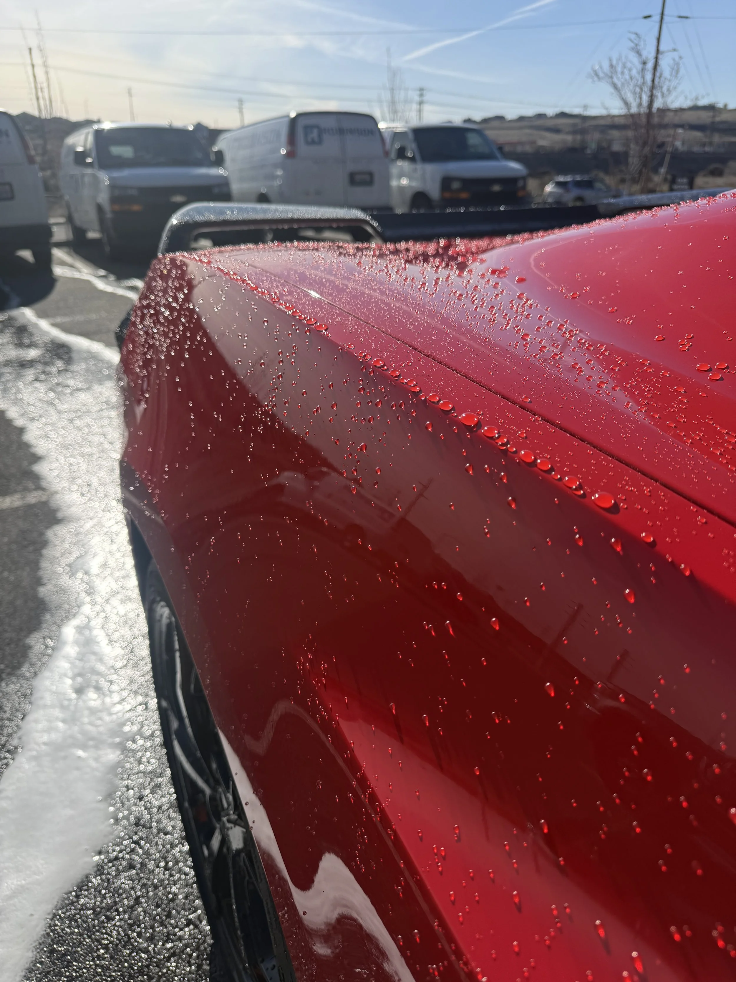 Close-up of a red car with water droplets on its surface, in a parking lot with white vans and a blue sky in the background.