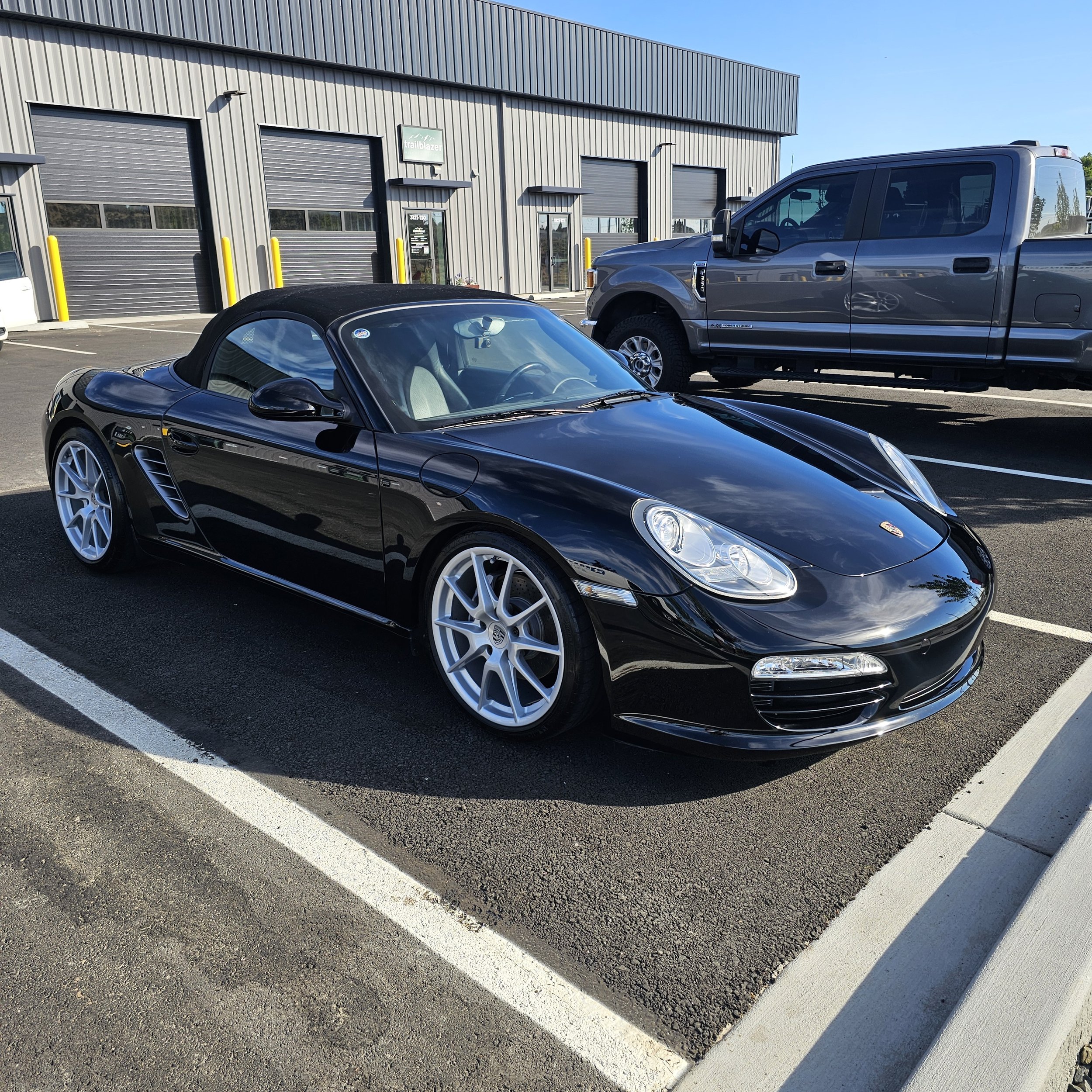 A parking lot in front of a gray industrial building with several garage doors and a sign that reads 'trailblazer'. There are three vehicles, including a black convertible sports car, a gray pickup truck, and a small white car. The sky is clear and sunny.