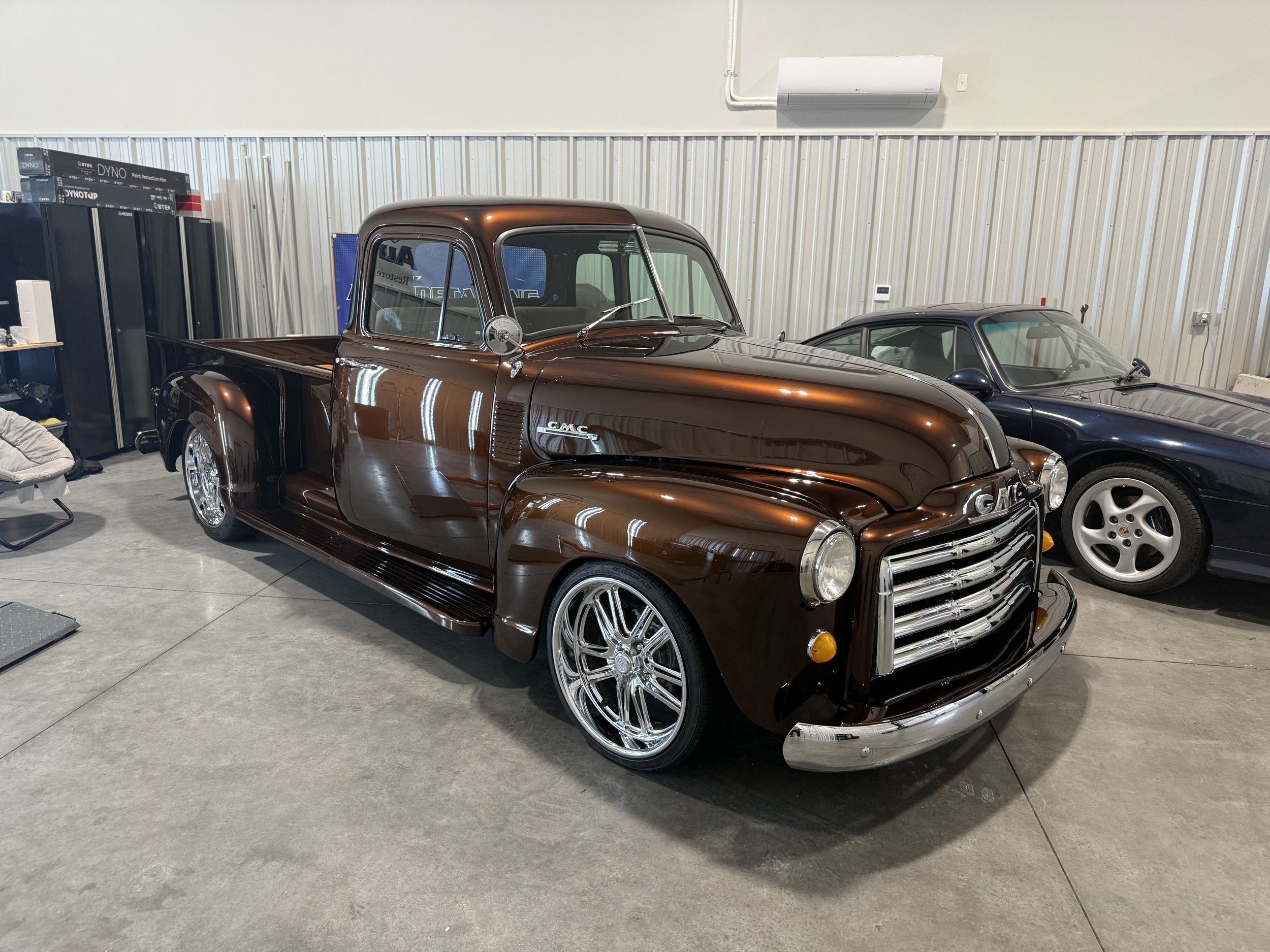 A vintage brown GMC pickup truck parked indoors next to a black sports car, with a metal wall, an air conditioning unit, and storage lockers in the background. Paint Correction