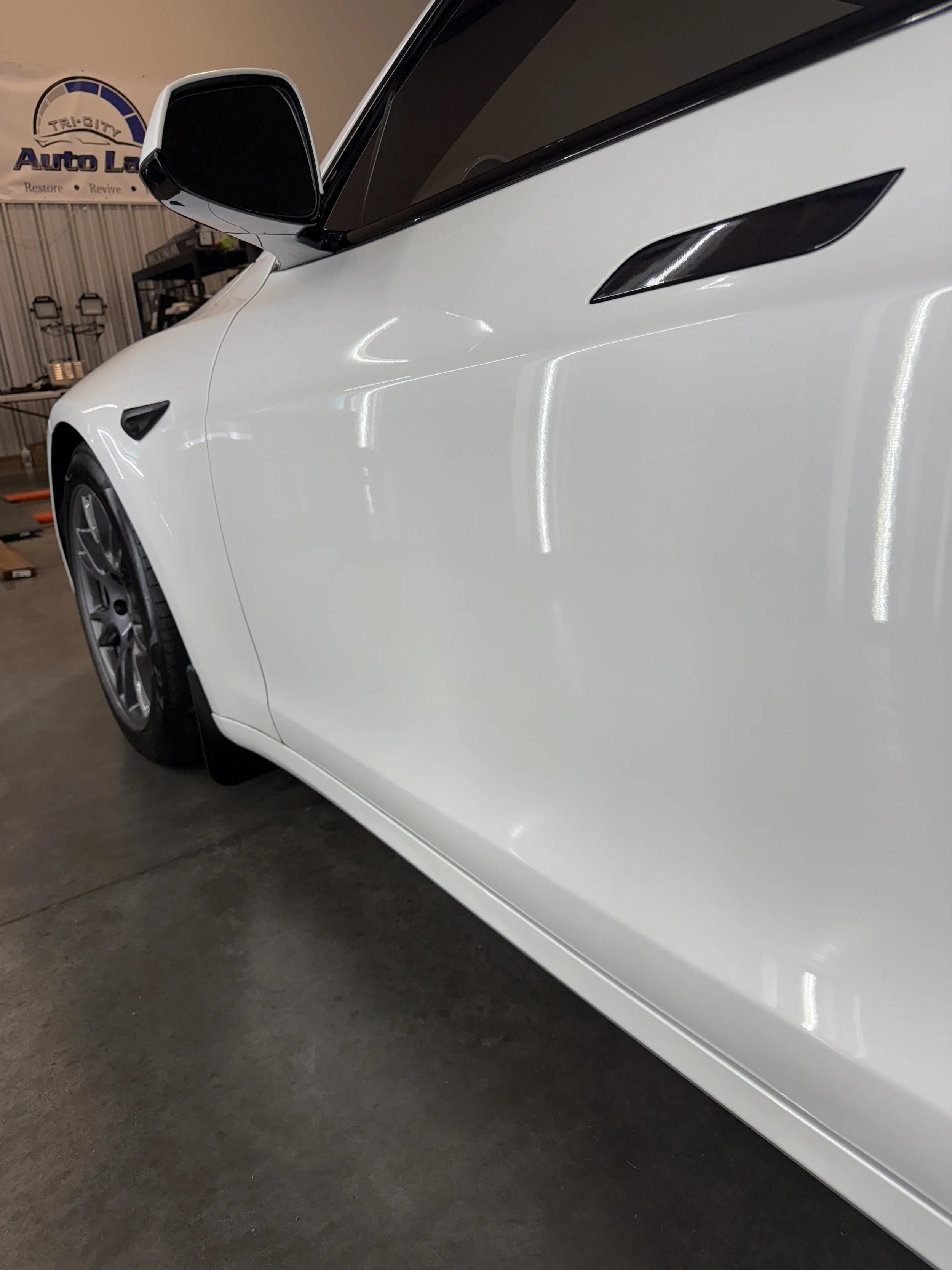 Close-up of a white car's front passenger side with a side mirror, tire, and headlight visible inside a garage. Ceramic Coating Gadsden, Alabama 
