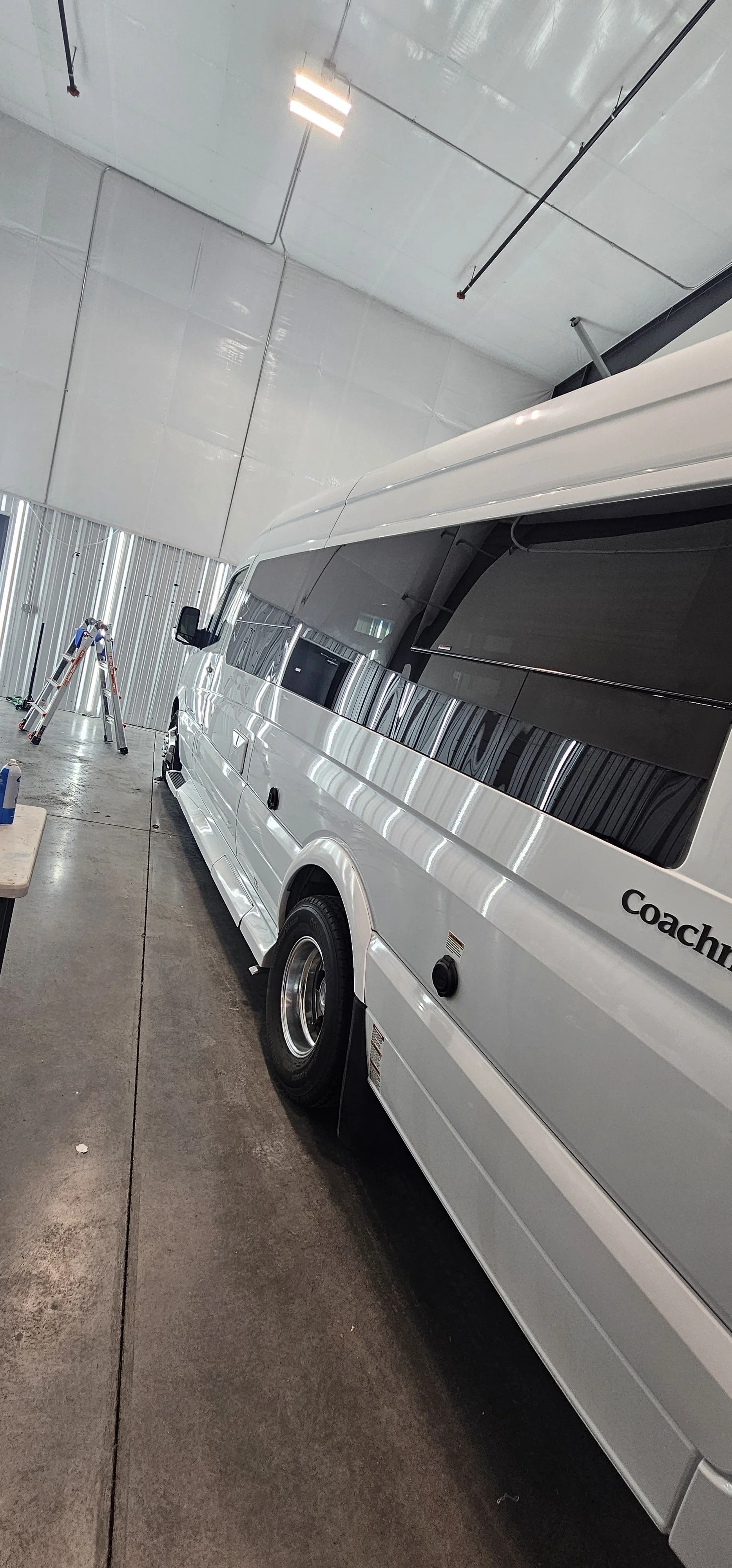 A white coach bus is parked inside a large industrial garage with a concrete floor and metal walls. There is a ladder in the background and a wooden workbench with bottled water on it. Paint Correction