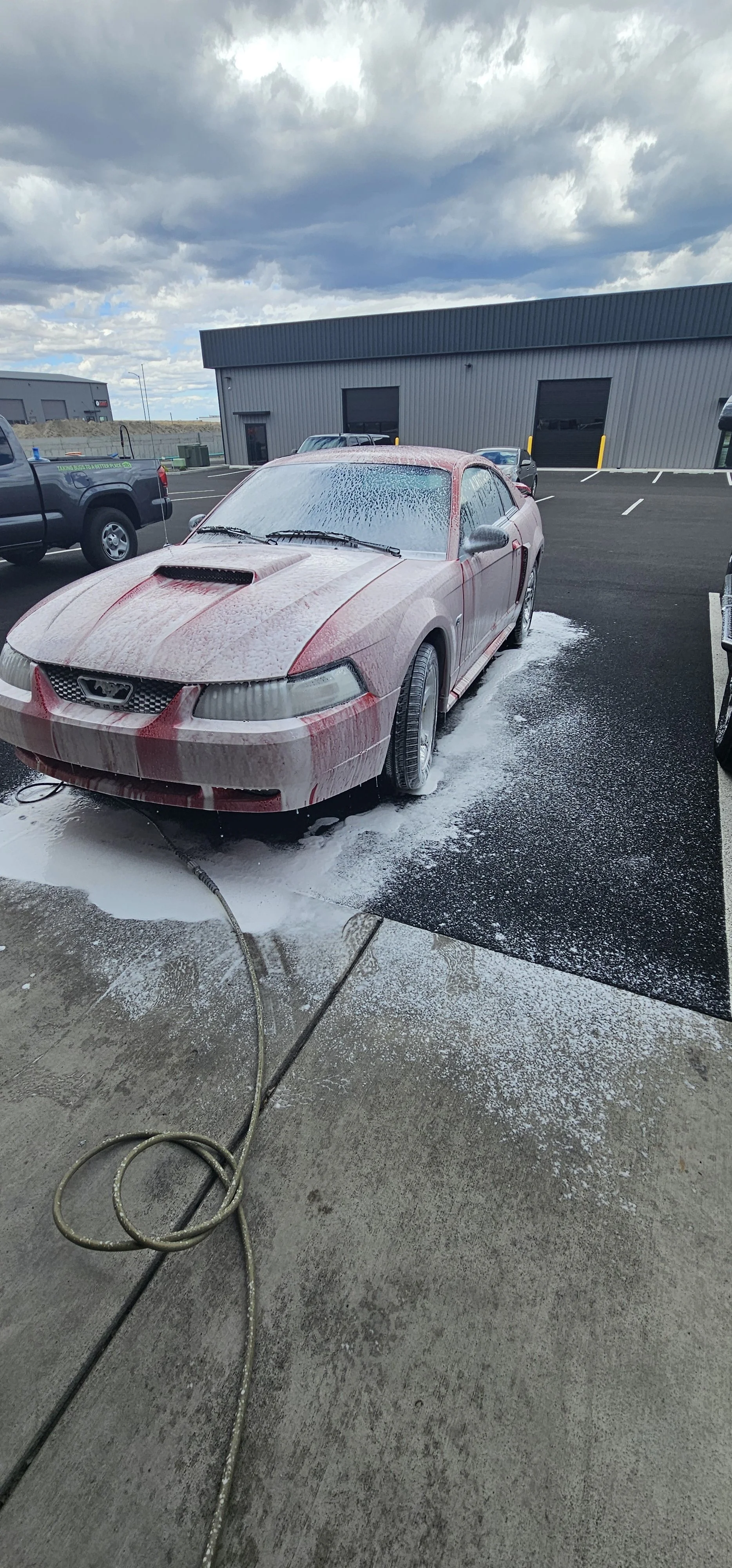 A red sports car is being washed with soap and water, sitting on a wet driveway with a waffle-weave soap suds covering the car, and a garden hose lying on the ground.
