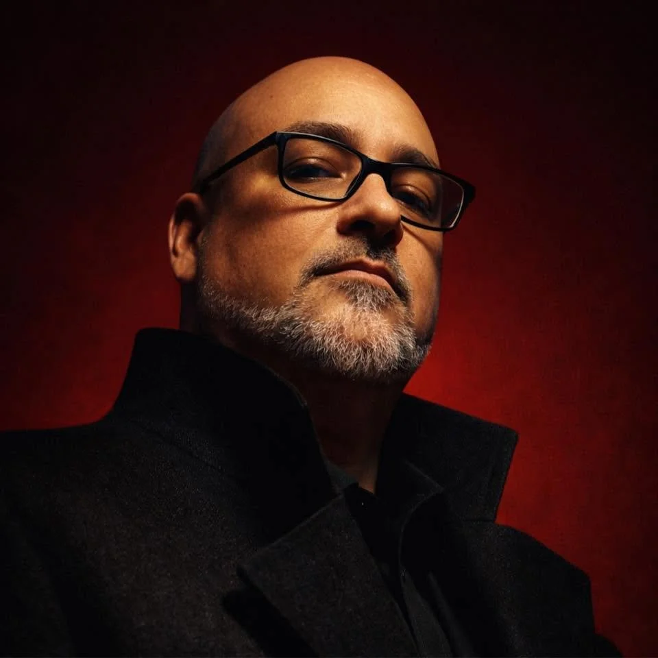 A bald man with glasses and a beard wearing a black coat, looking confidently at the camera against a dark red background. Kyle McIntosh. Deck cuts. Artist.