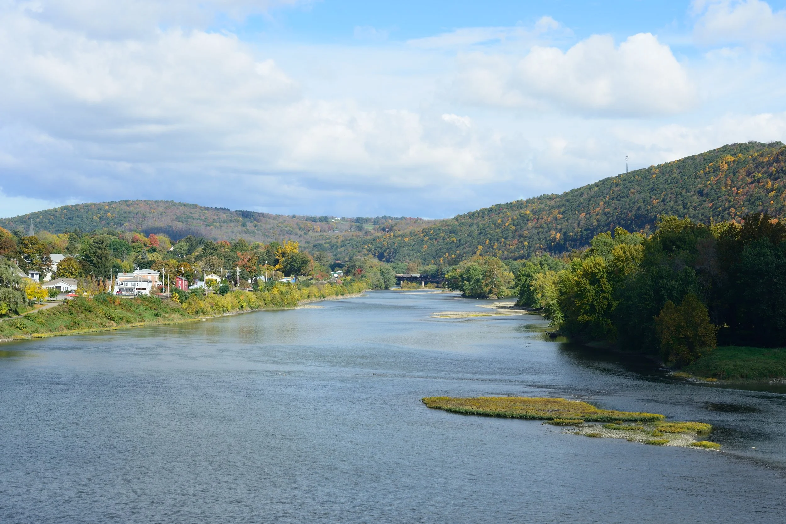 A river flowing through a landscape with trees on both sides and hills in the background under a partly cloudy sky.