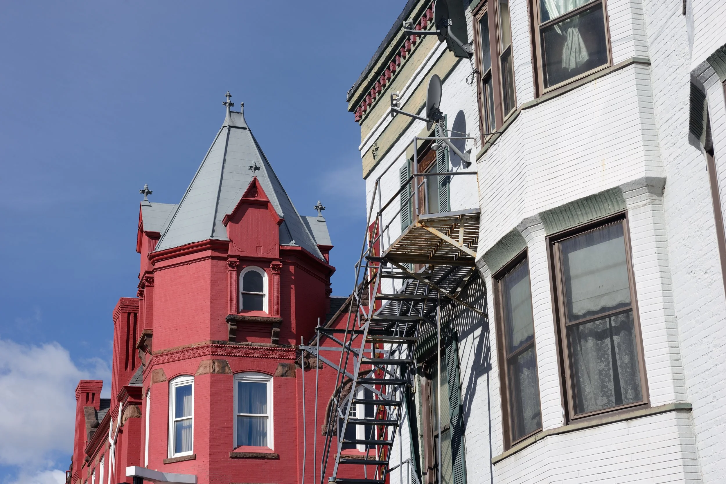 A red Victorian-style building with decorative trim and multiple windows topped with a pointed turret, alongside a white building with green window frames and an external fire escape staircase, under a blue sky with a few clouds.