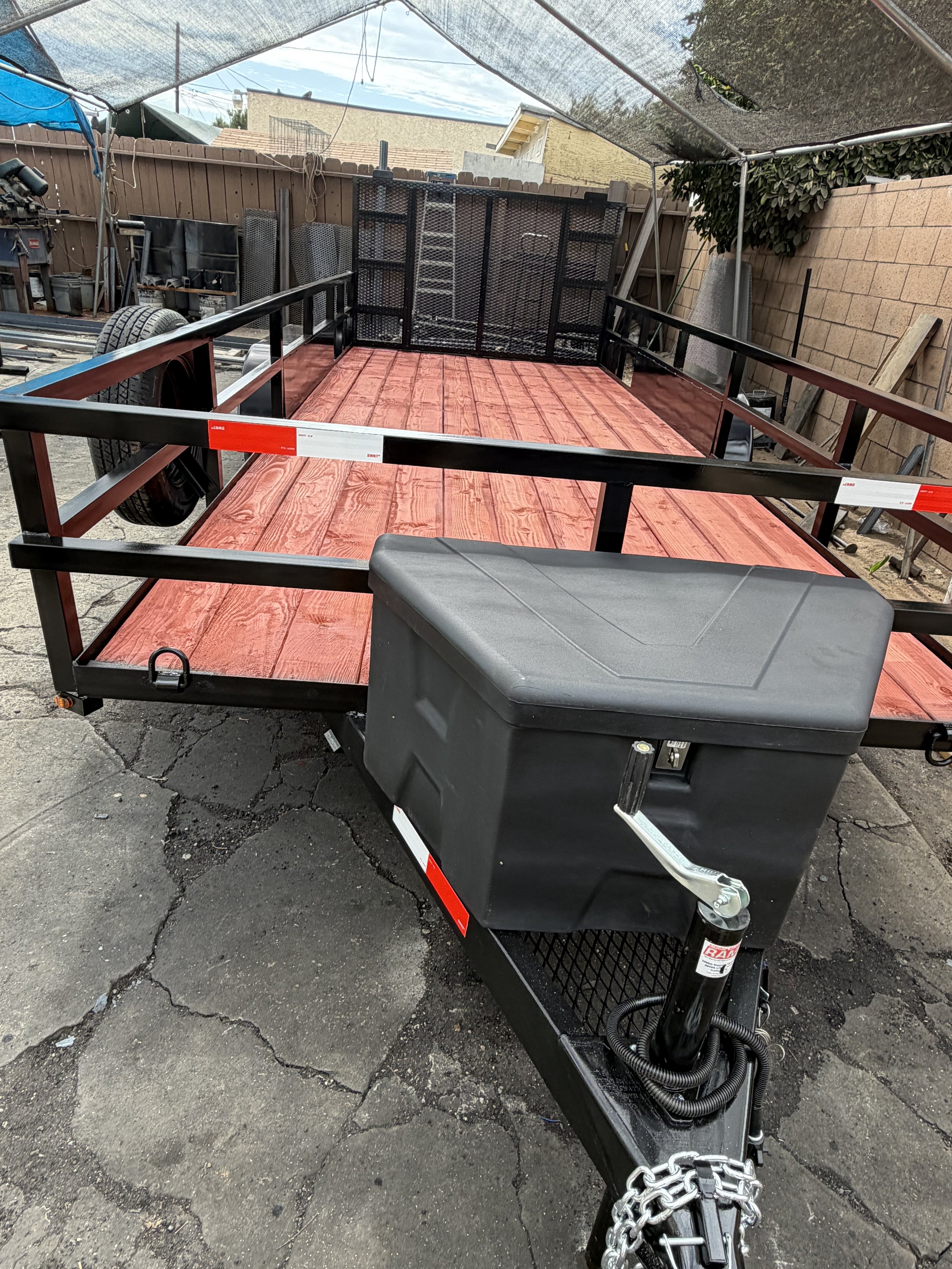 A flatbed utility trailer with a black metal frame and red wood floor in a backyard, with a chain and hitch at the front, and a black storage container on the ground nearby.
