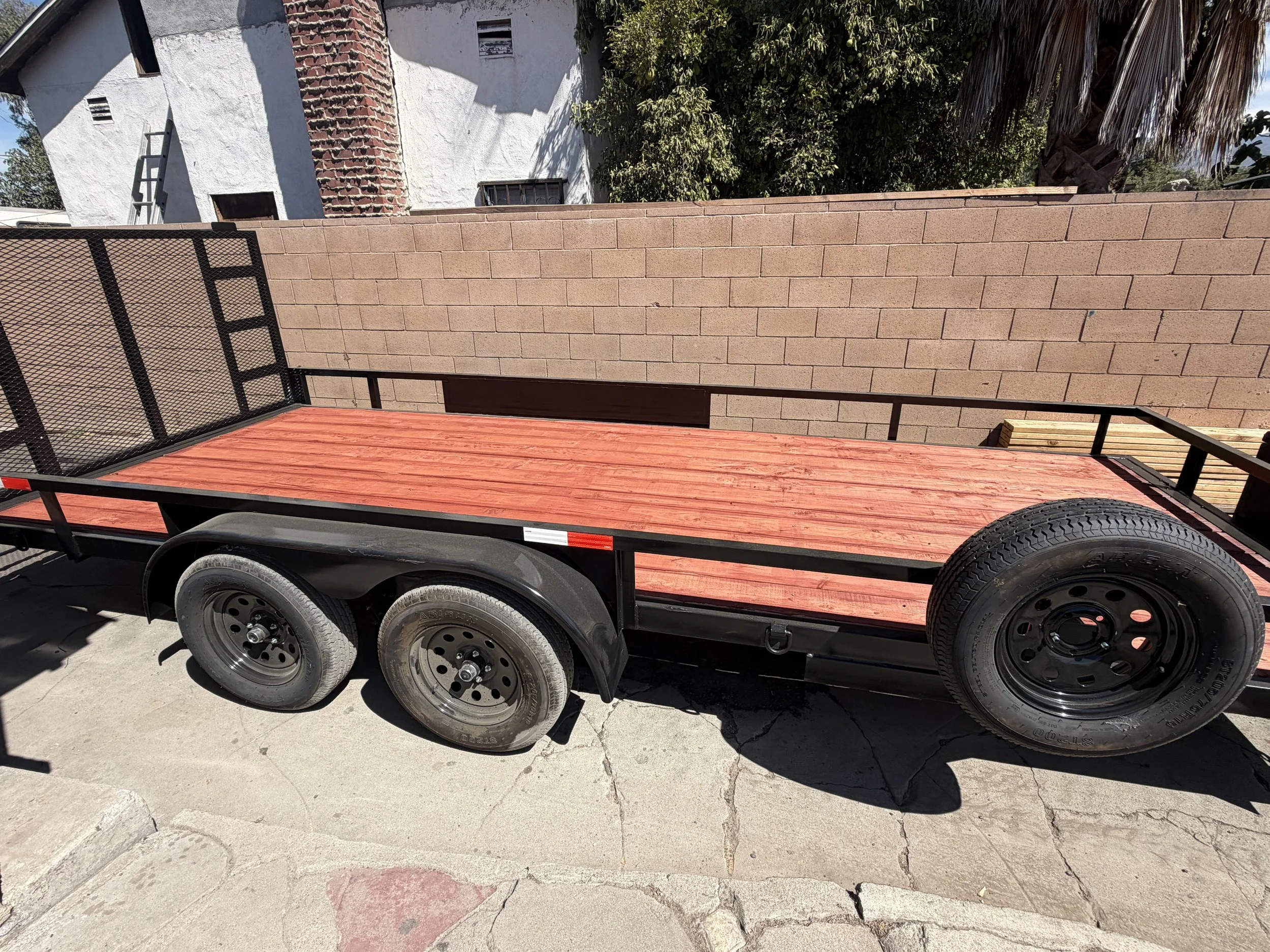 Image of a flatbed trailer with black tires and a red wooden deck, parked on a concrete surface. A brick and cement wall is visible in the background with some greenery and a white building.