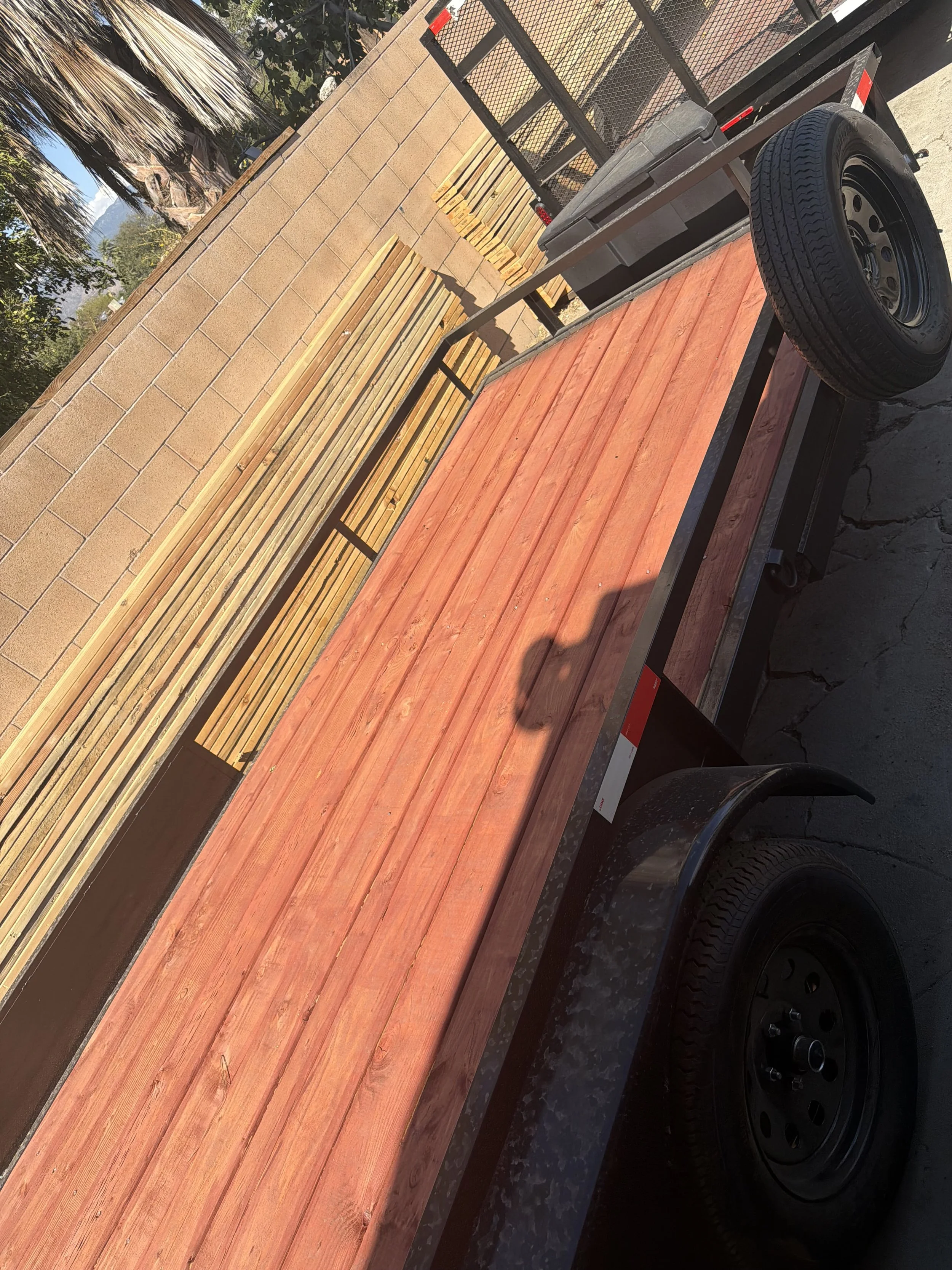 A flatbed trailer loaded with wooden planks parked outdoors, with a brick wall and trees in the background, and the sky partly cloudy.