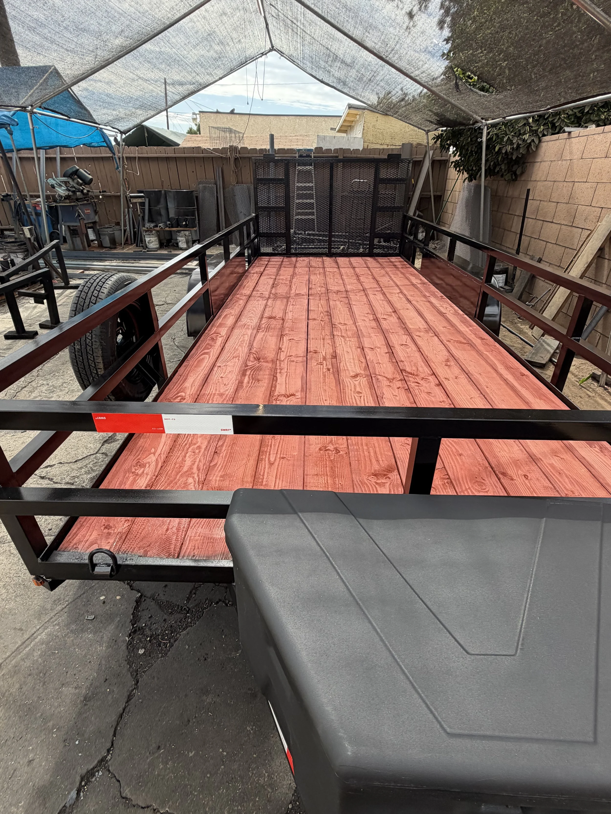 A flatbed utility trailer with a wooden floor and black metal frame, parked on a cracked asphalt surface. The trailer has black wheels with tires and a gray plastic storage box at the front. There's a lens reflection at the bottom, and in the background, there are black metal shelves, a fence, and parts of neighboring buildings.