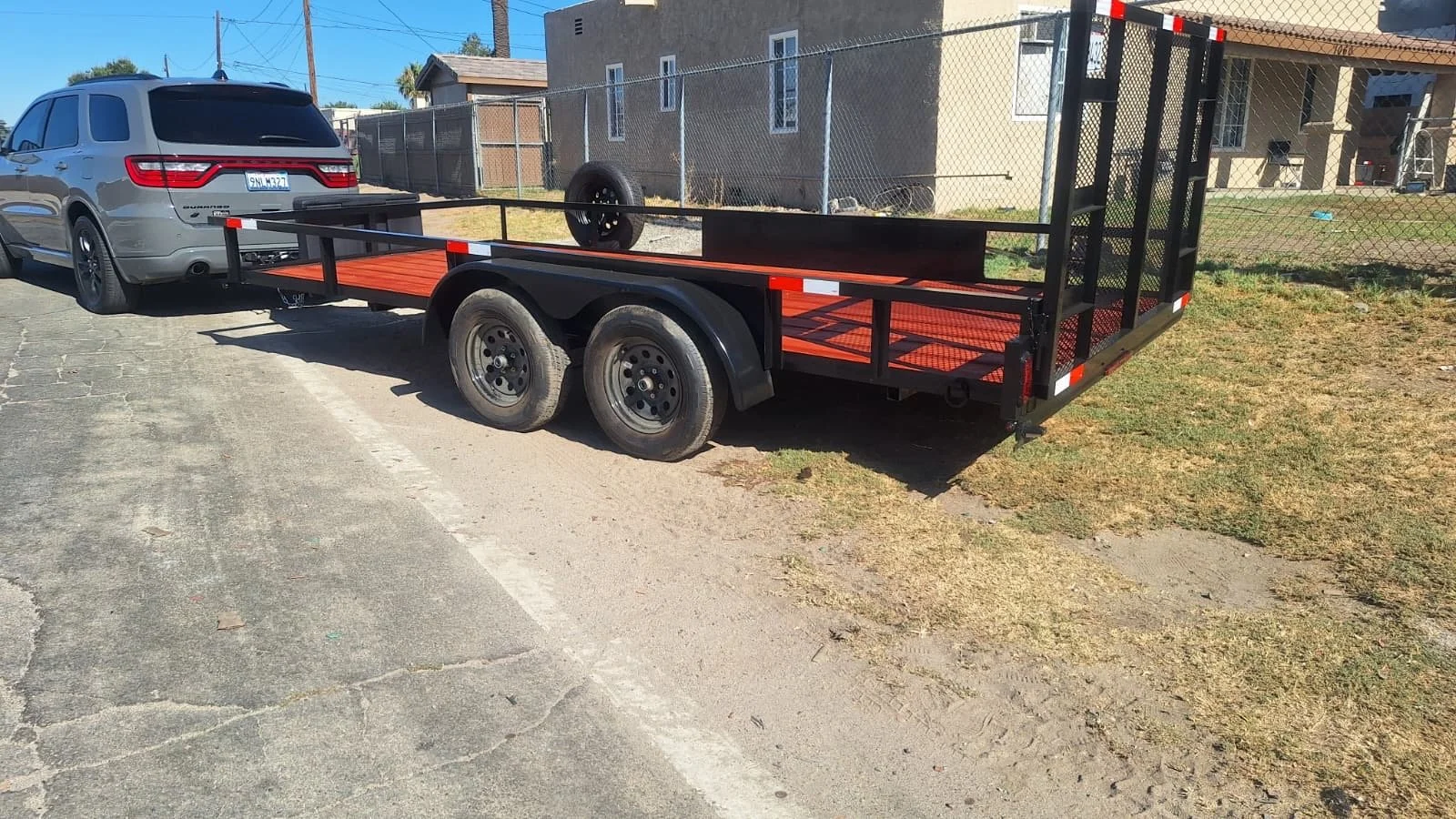 A black flatbed utility trailer attached to a parked silver SUV in a residential area, with a chain-link fence and houses in the background.