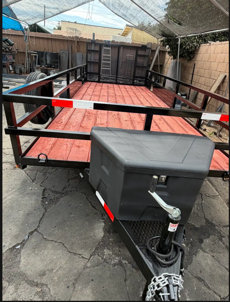 A flatbed trailer with a red wooden floor, black metal rails, and a black toolbox at the front, parked on cracked pavement in an outdoor yard.
