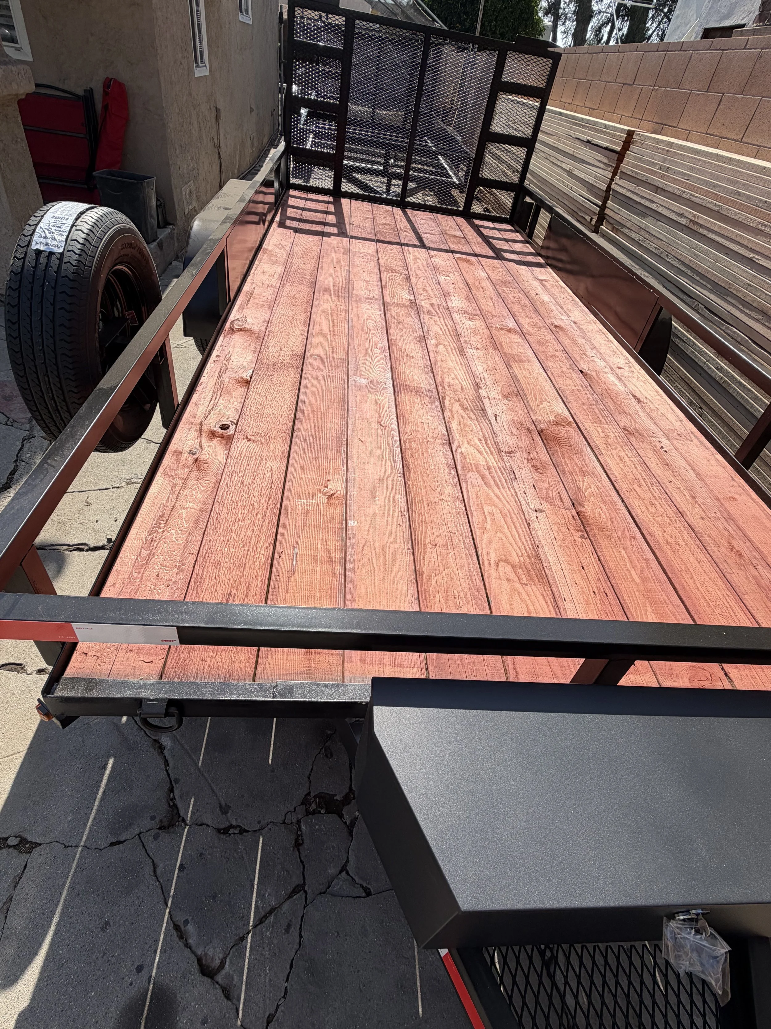 A red wooden flatbed trailer with black metal frame and sides, parked on cracked asphalt ground, with stacked bricks and a brick wall in the background.