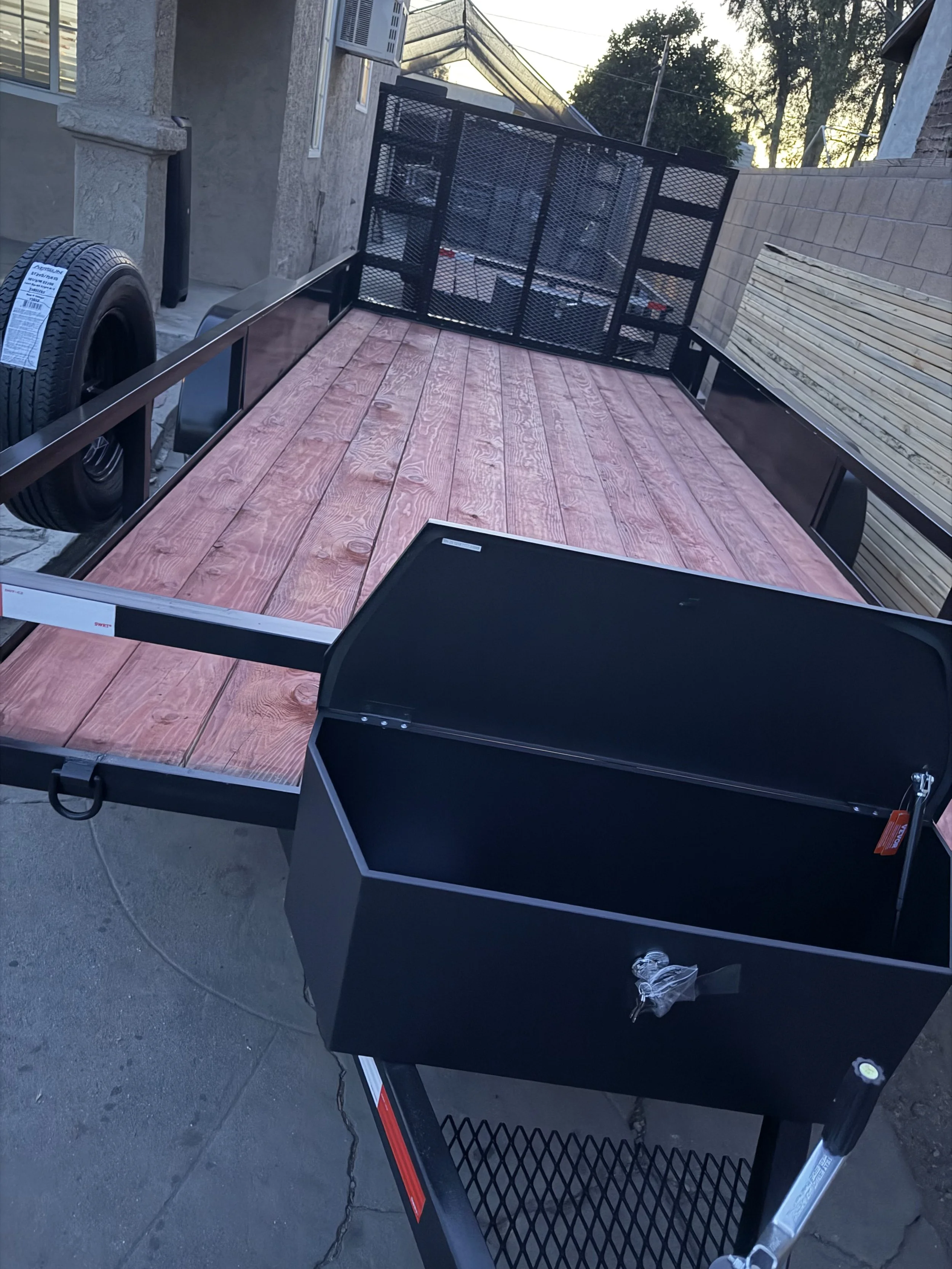 A black utility trailer with a pink wooden bed parked on concrete pavement; the trailer has a metal side panel and a lifted tailgate, and a spare tire attached to its front side.