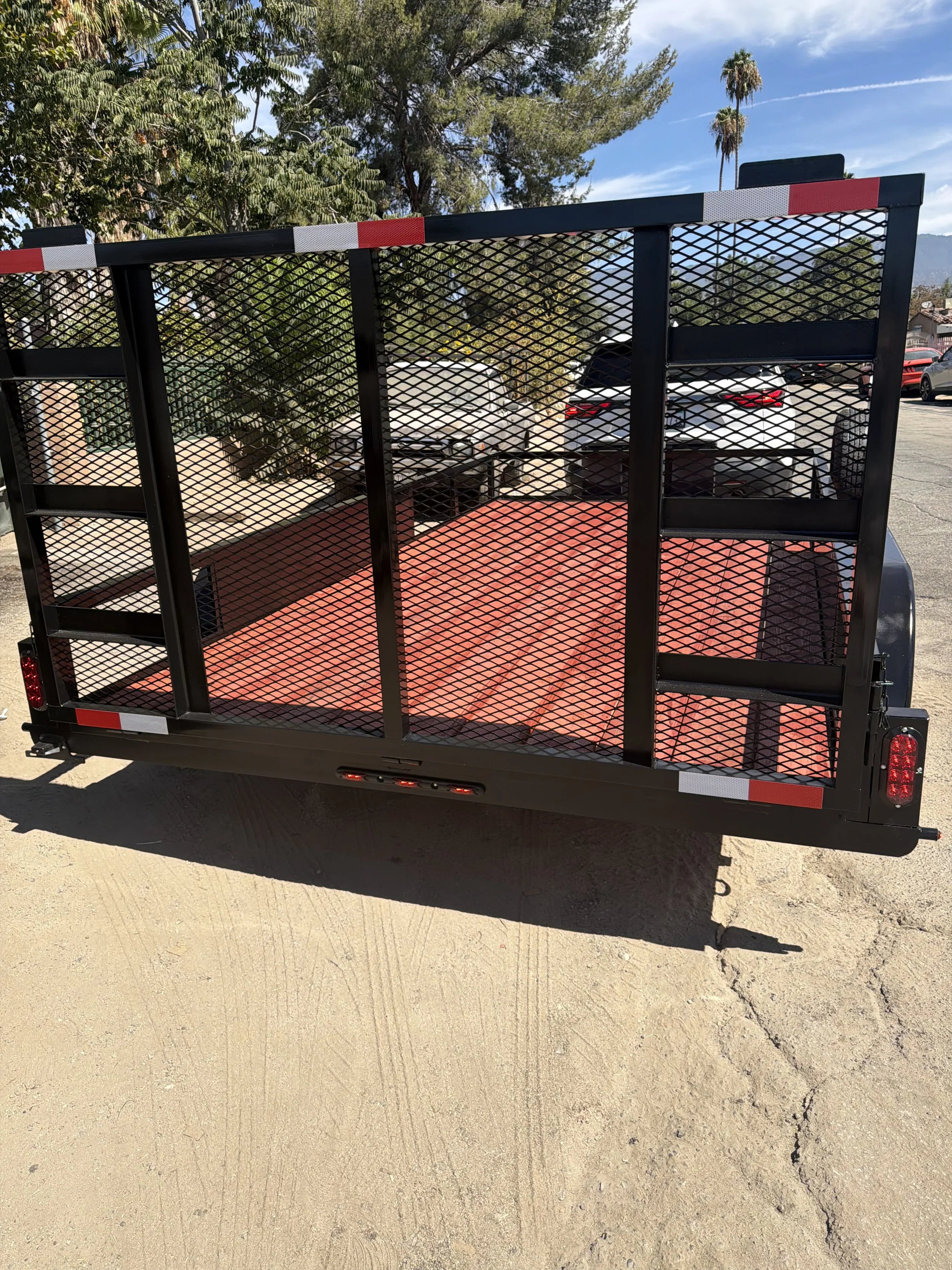 Black utility trailer with red flooring and black metal fencing parked on a dirt lot with trees and a blue sky in the background.